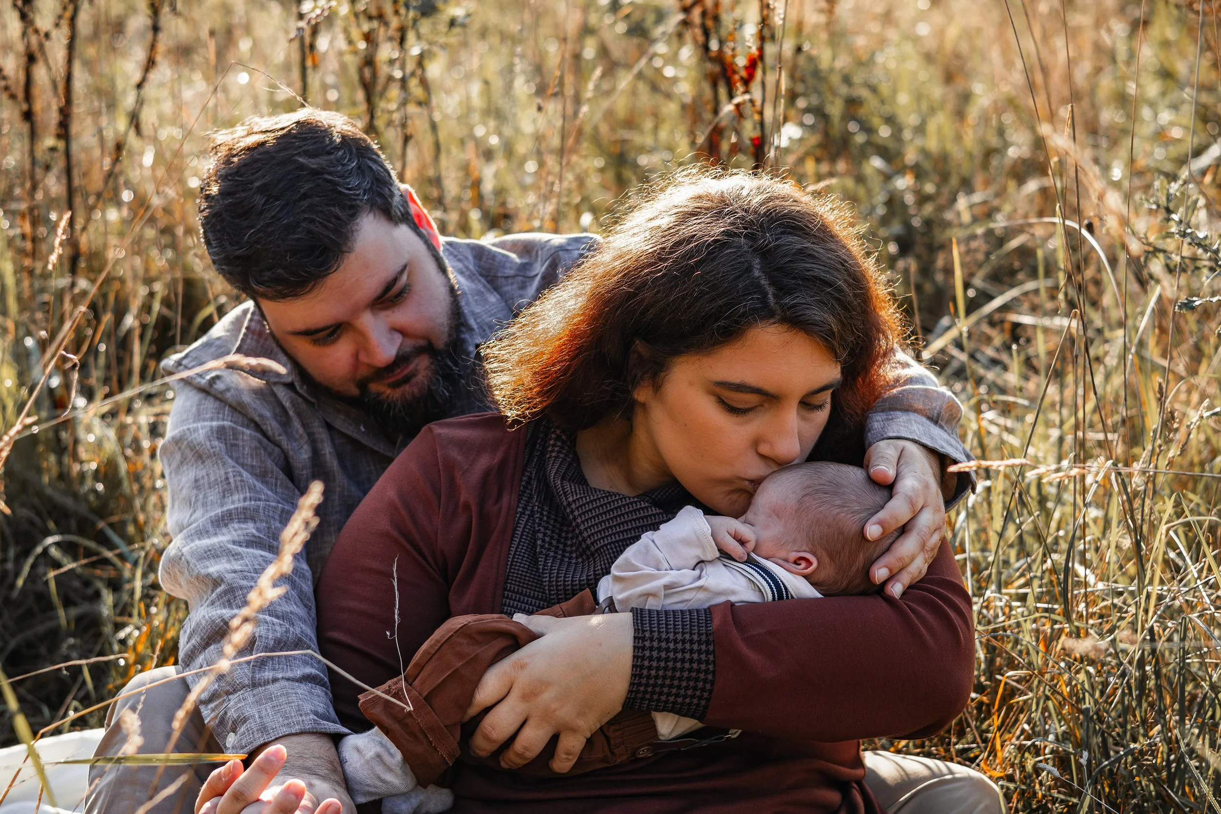 Ein Mann und eine Frau sitzen in einem Feld, der Mann umarmt die Frau von hinten während sie ein Baby hält und küsst.
