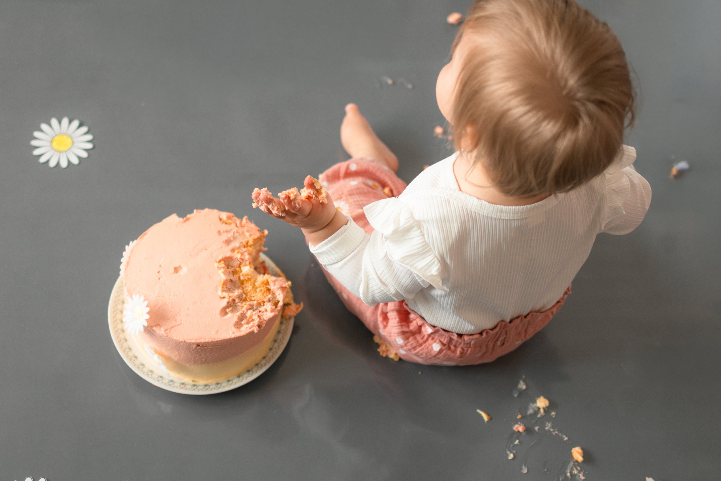 Kleines Kind sitzt auf dem Boden und isst einen Kuchen, der teilweise gegessen ist. Der Kuchen liegt auf einem Teller, der auf einem schwarzen Boden platziert ist. Es gibt auch einige verstreute Kekskrümel und dekorative Papiere, die mit Blumenmotive