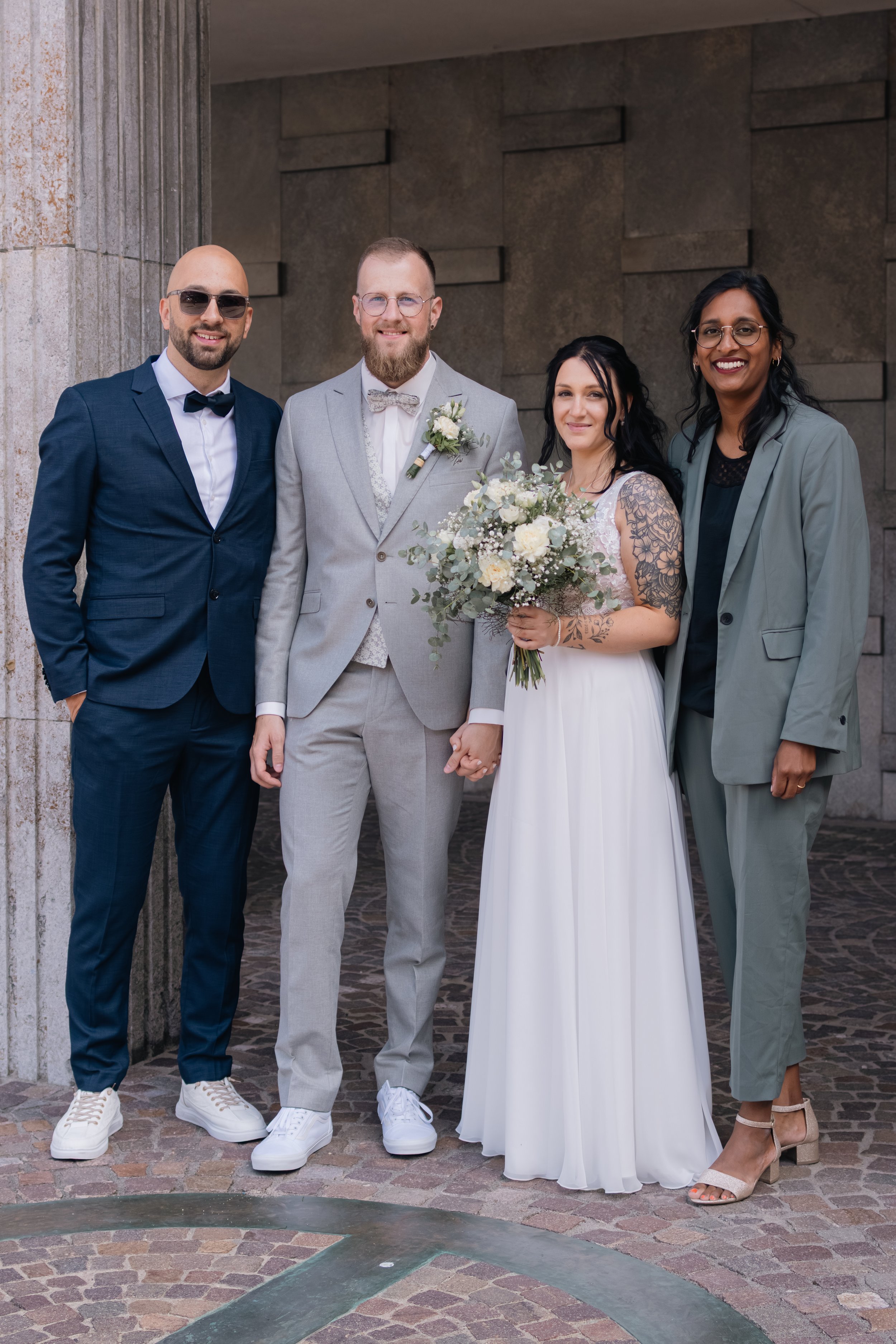 Gruppe von vier Menschen bei einer Hochzeit, bestehend aus einem Bräutigam in grauer Anzug, einer Braut in weißem Kleid mit Blumenstrauß, und zwei weitere Personen in schicken Anzügen, vor einer Steinmauer.