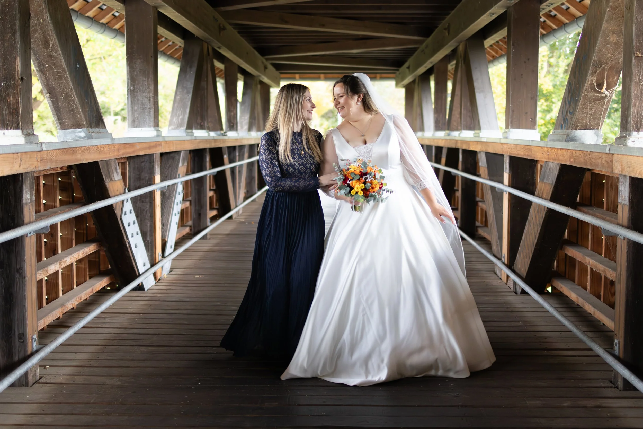 Zwei Frauen, eine in einem Hochzeitskleid mit Brautstrauß, und die andere in einem schwarzen Kleid, stehen sich auf einer Holzbrücke gegenüber und lachen, umgeben von einem natürlichen Hintergrund.