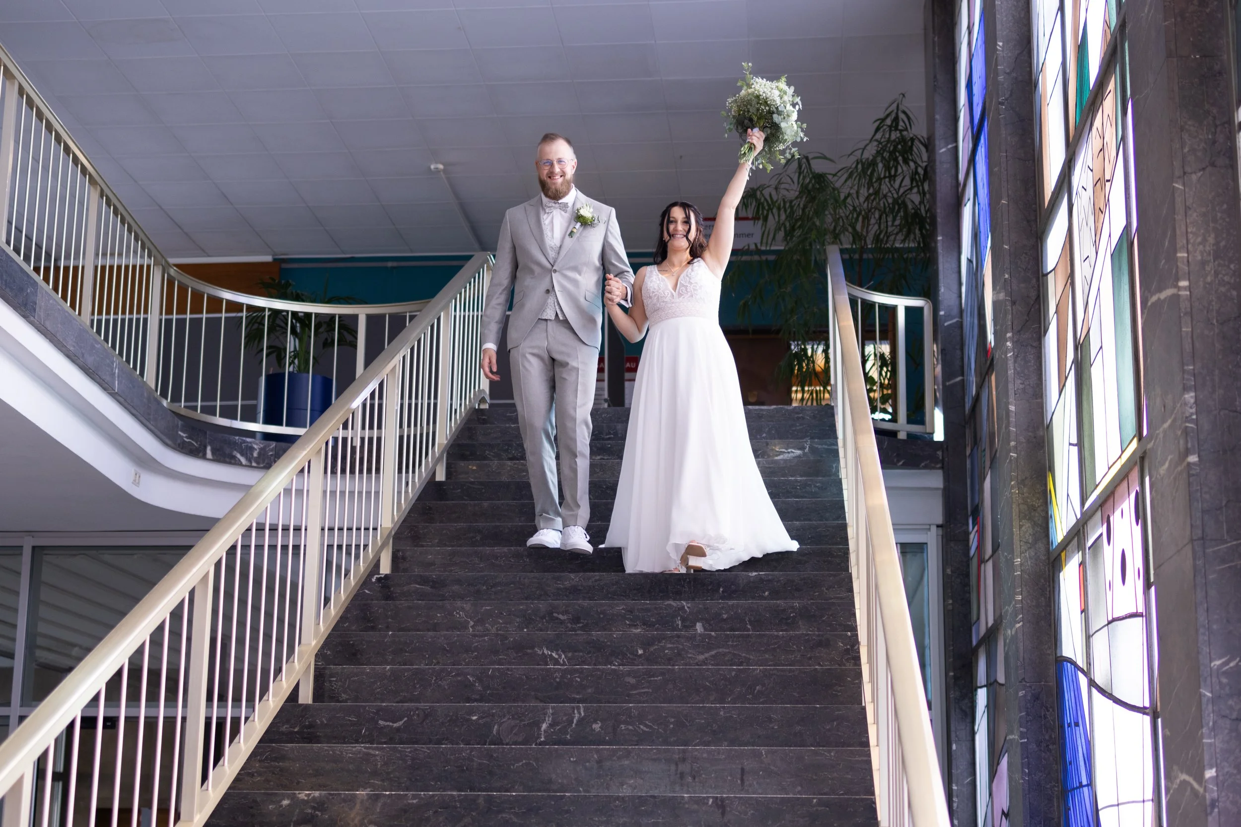 Ein Brautpaar auf einer Treppe, die Braut hält einen Blumenstrauß in der Hand und freut sich, während er neben ihr steht, beide tragen formelle Hochzeitskleidung.