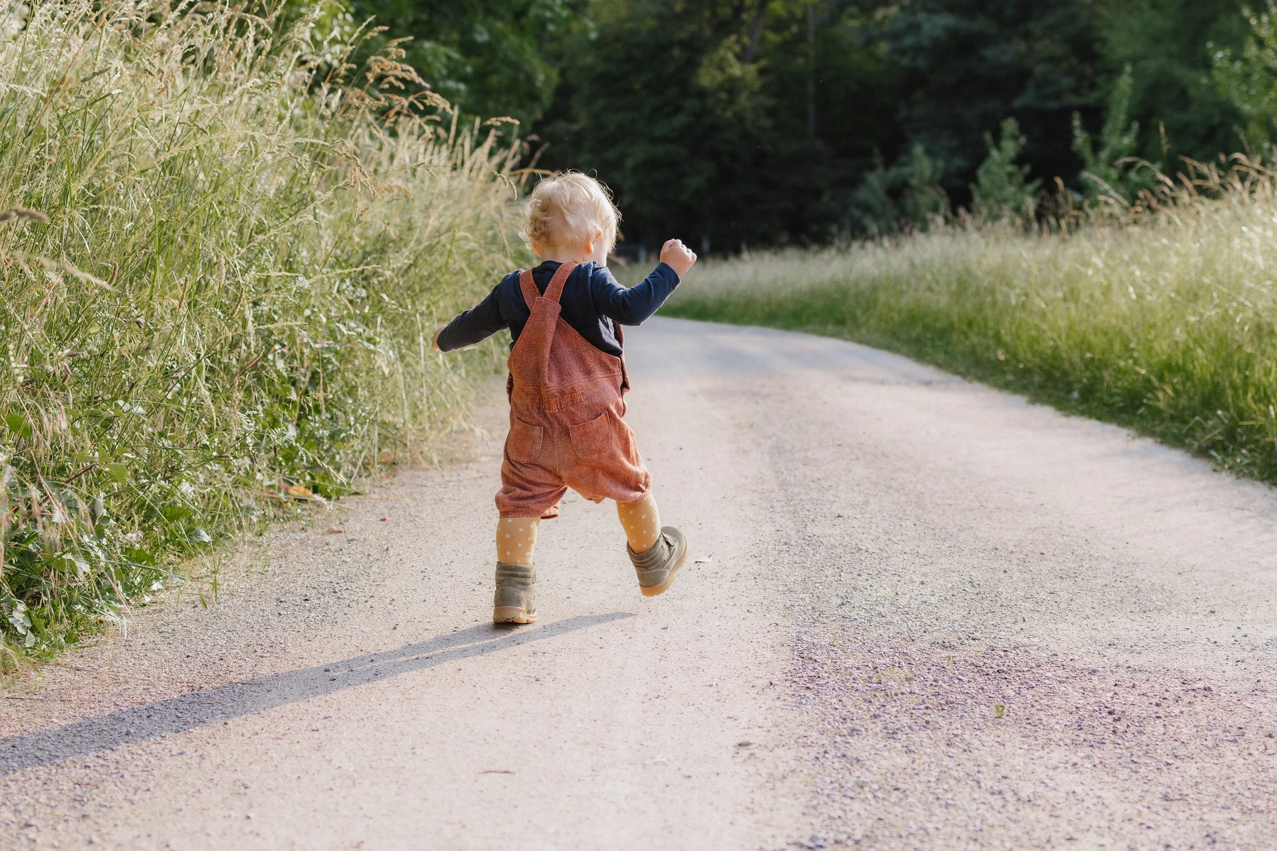 Ein kleines Kind mit einem roten Rucksack läuft auf einer asphaltierten Landstraße durch eine grüne Naturumgebung, wahrscheinlich im Freien, bei Sonnenlicht.