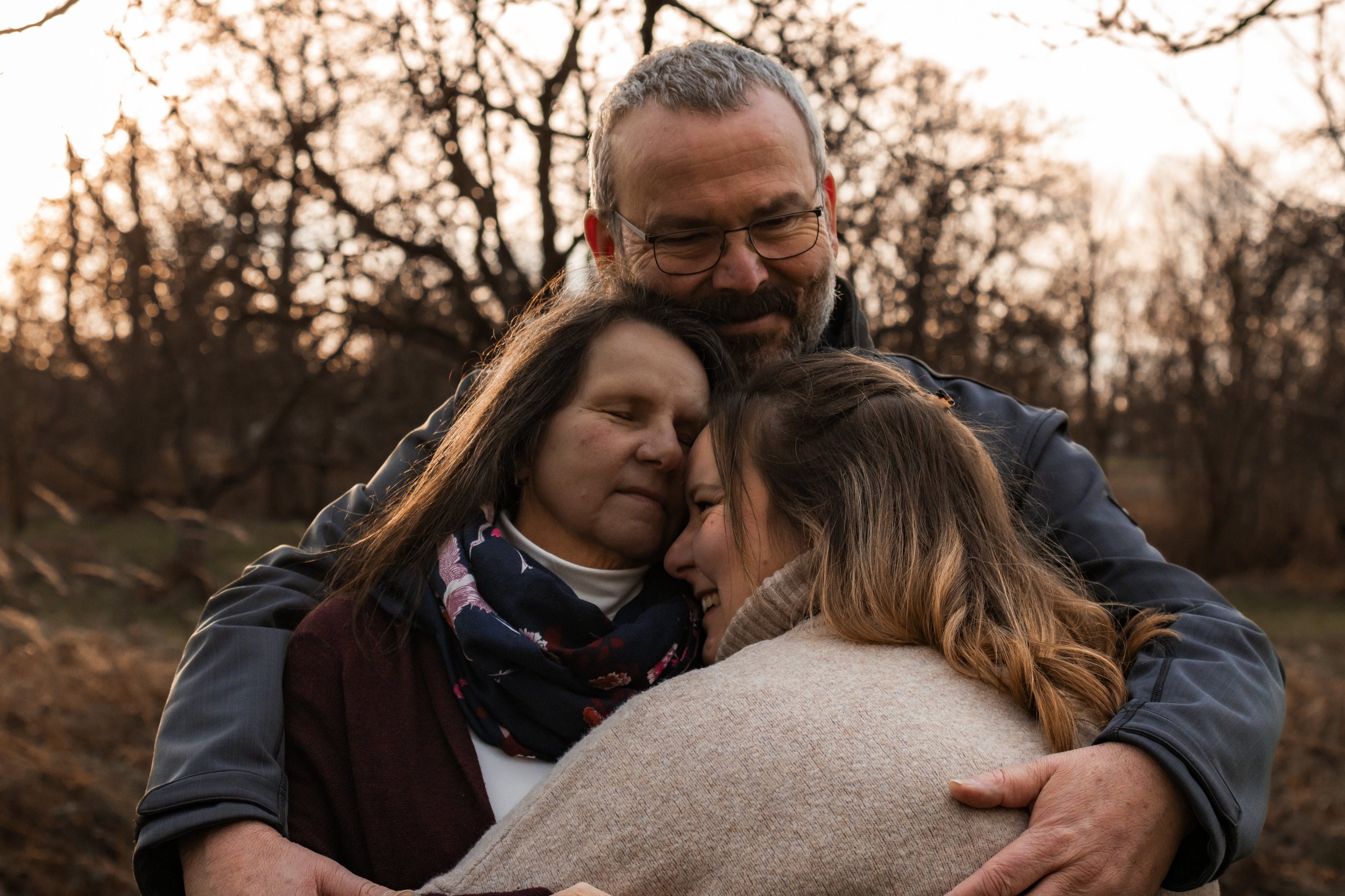 Drei Menschen umarmen sich innig im Freien bei Sonnenuntergang, umgeben von Bäumen ohne Blätter.
