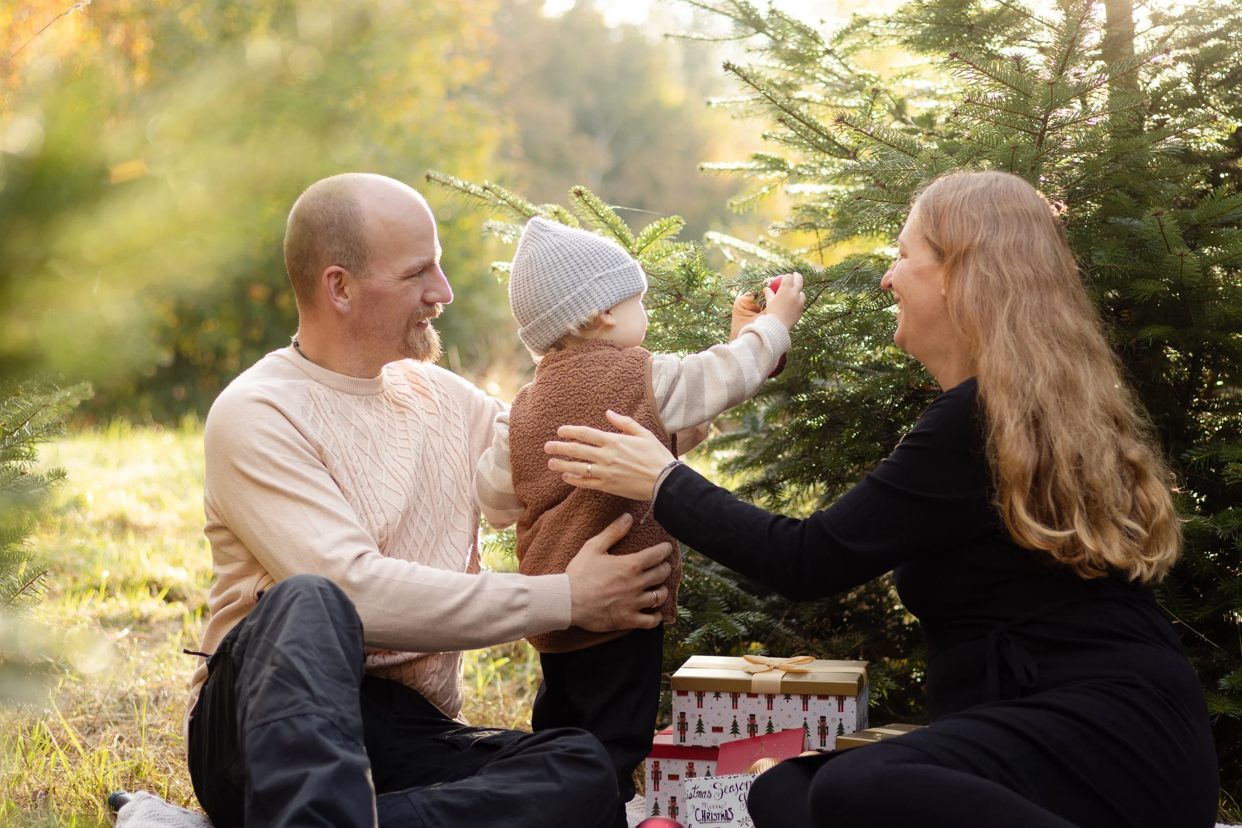 Familie sitzt im Freien und schmückt einen Weihnachtsbaum, mit Geschenken darunter.