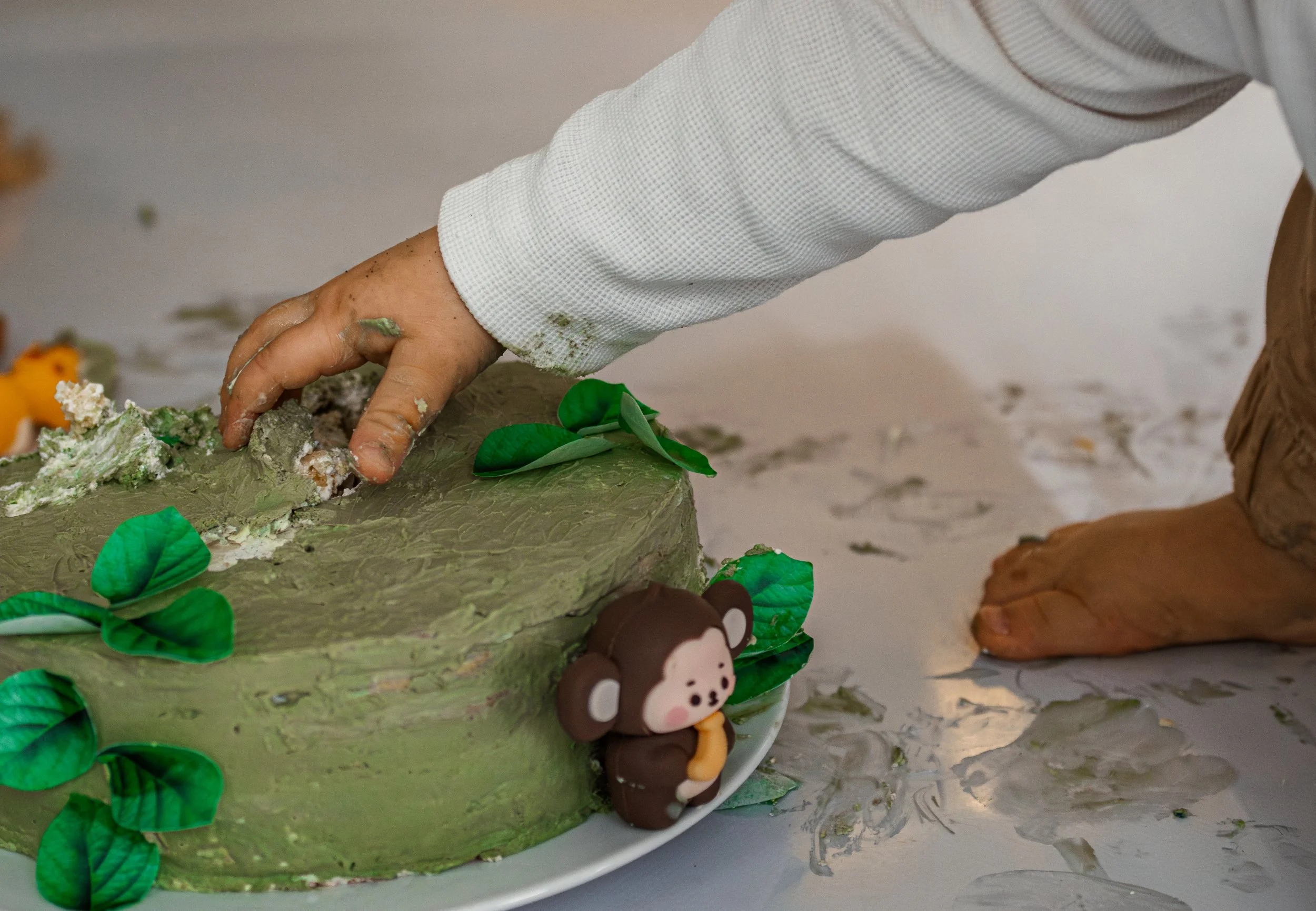 Eine Kinderhand dekoriert einen Dinosaurier-Kuchen mit grüner Dekoration. Ein Teddybär-Schneemann steht neben dem Kuchen auf einem weißen Teller.