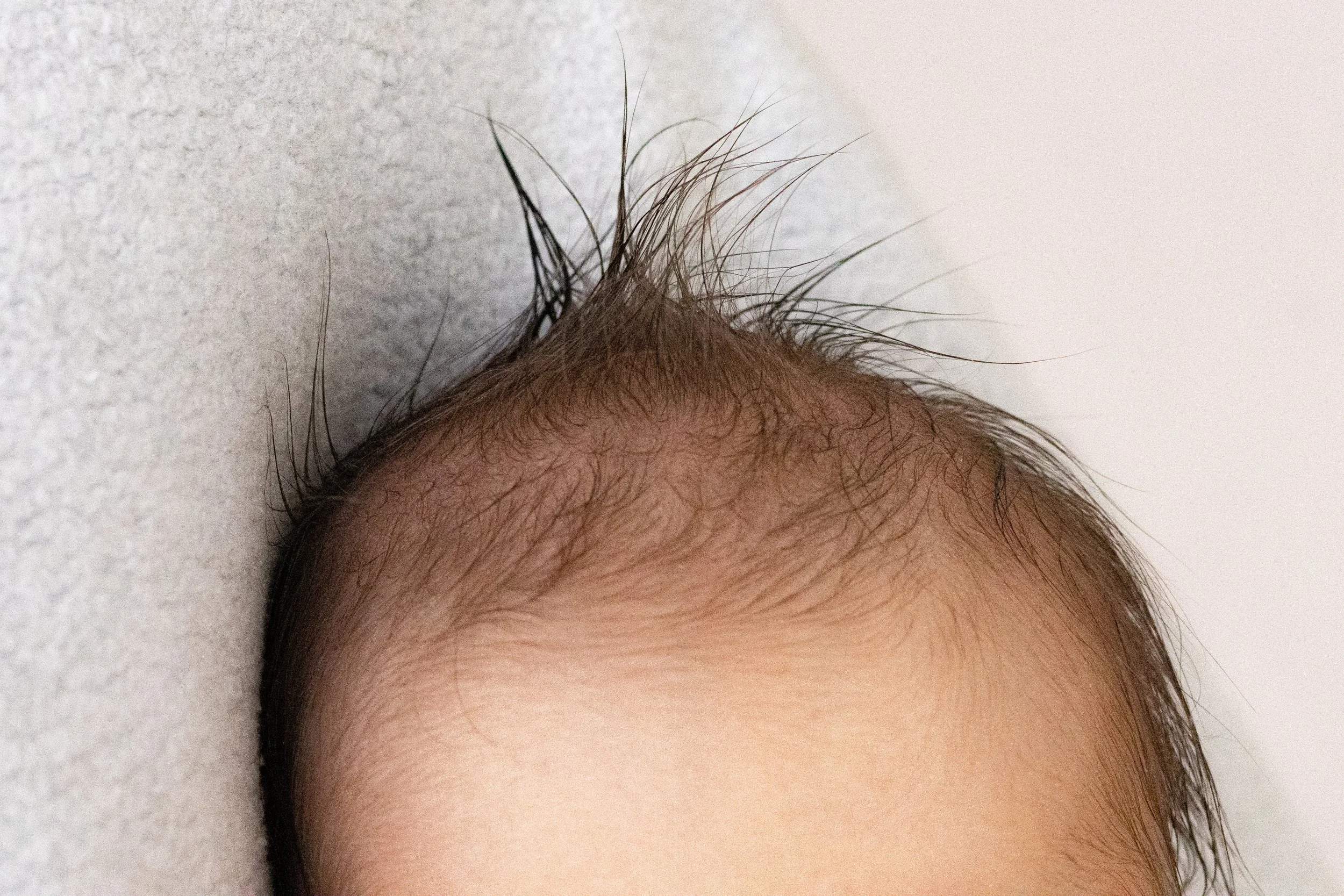 Nahaufnahme eines Babykopfes mit dünnem, spärlichem Haar, liegend auf einem weichen, hellgrauen Untergrund.