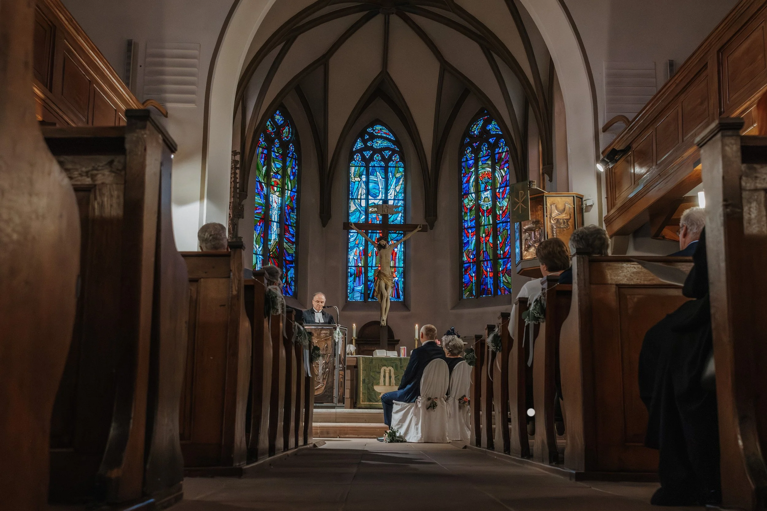 Eine Hochzeit in einer Kirche, mit Paaren, die vor dem Altar sitzen, einem Priester, der eine Zeremonie durchführt, und bunten Buntglasfenstern im Hintergrund.