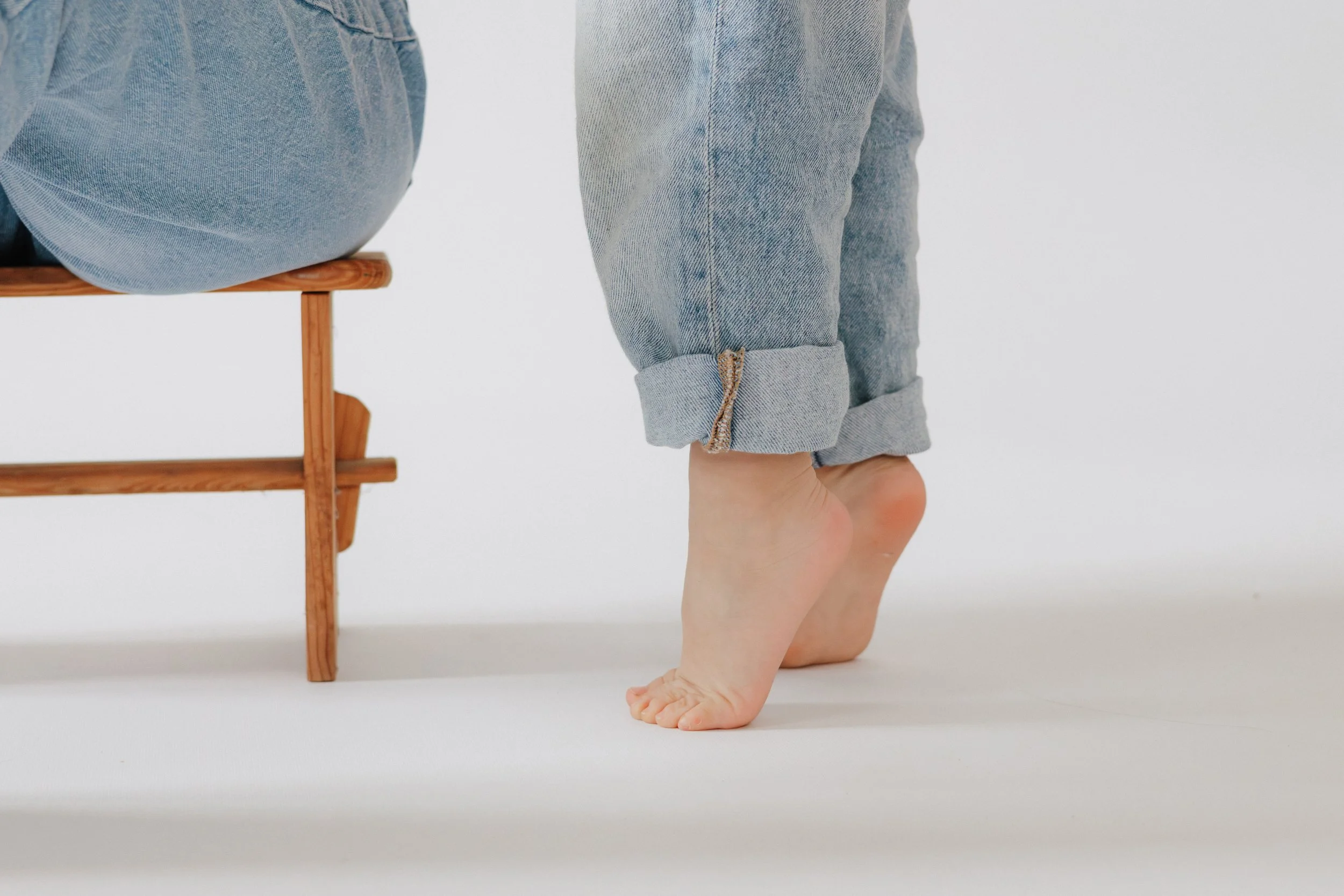 Ein Kind kniet auf dem Boden, trägt Jeans und ist nur teilweise im Bild sichtbar. Es befindet sich neben einem kleinen Holzstuhl gegen einen weißen Hintergrund.