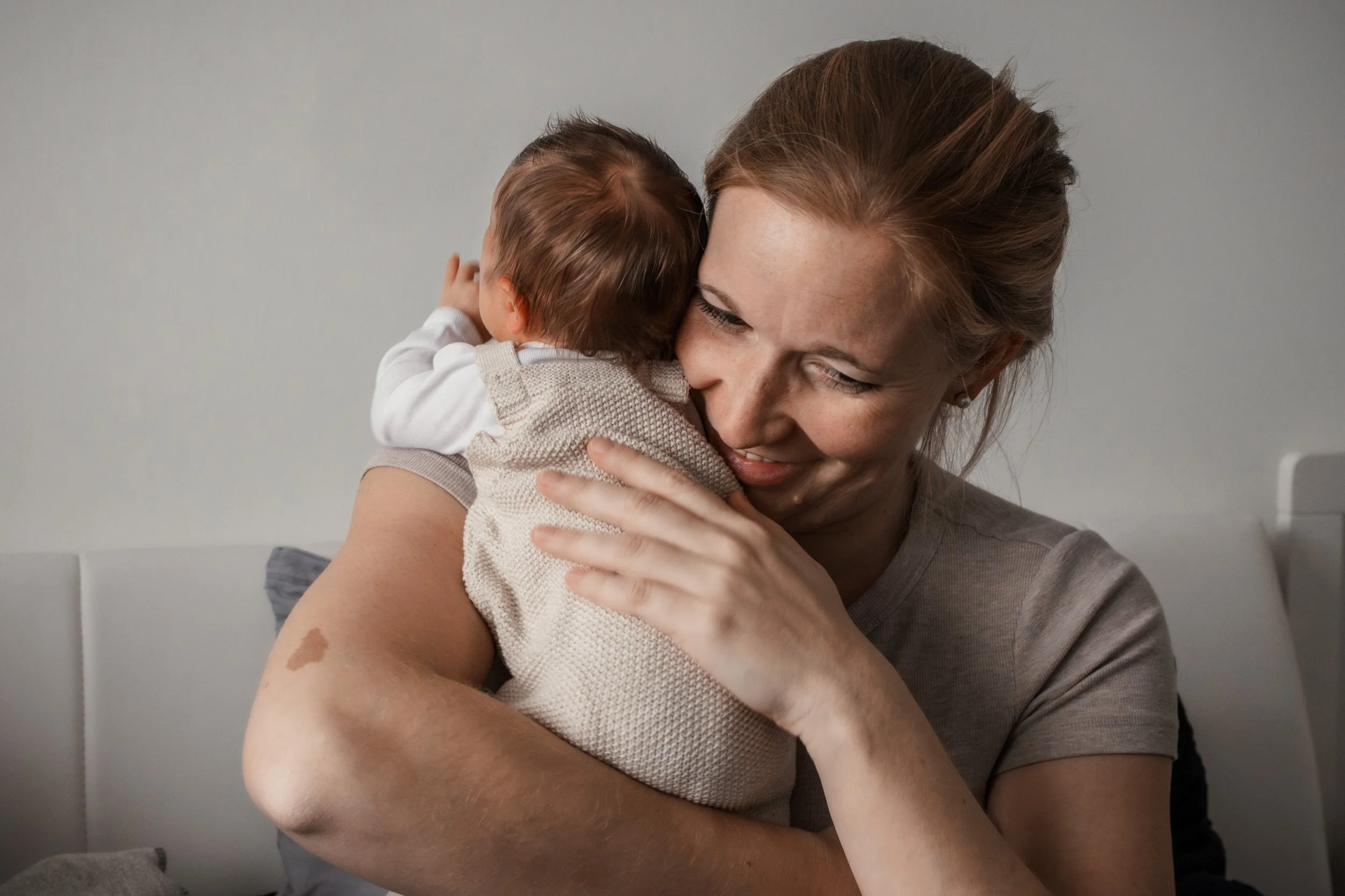 Eine Frau hält und umarmt ein Baby, das ihm ihr Gesicht an das ihrer Schulter legt, in einem Zimmer mit neutralem Hintergrund.