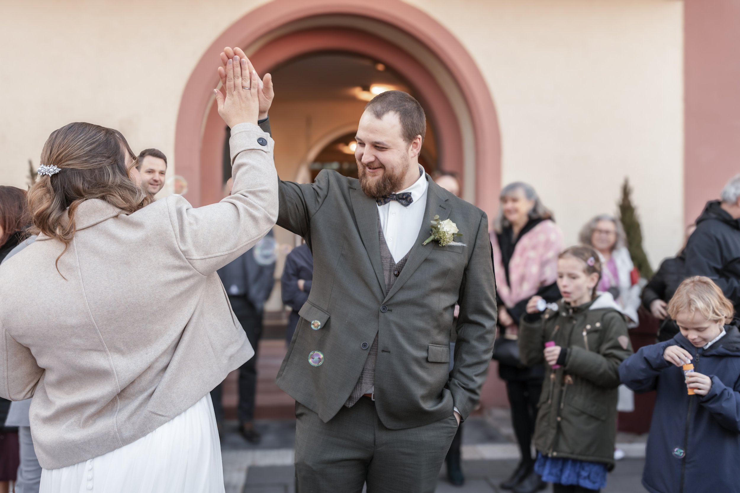 Ein Mann und eine Frau tauschen bei einer Feier eine High-Five, umarmt von mehreren Gästen im Hintergrund. Der Mann trägt einen schwarzen Anzug mit Fliege, die Frau trägt einen beigen Mantel. Es findet wahrscheinlich eine Hochzeit oder ein festliches