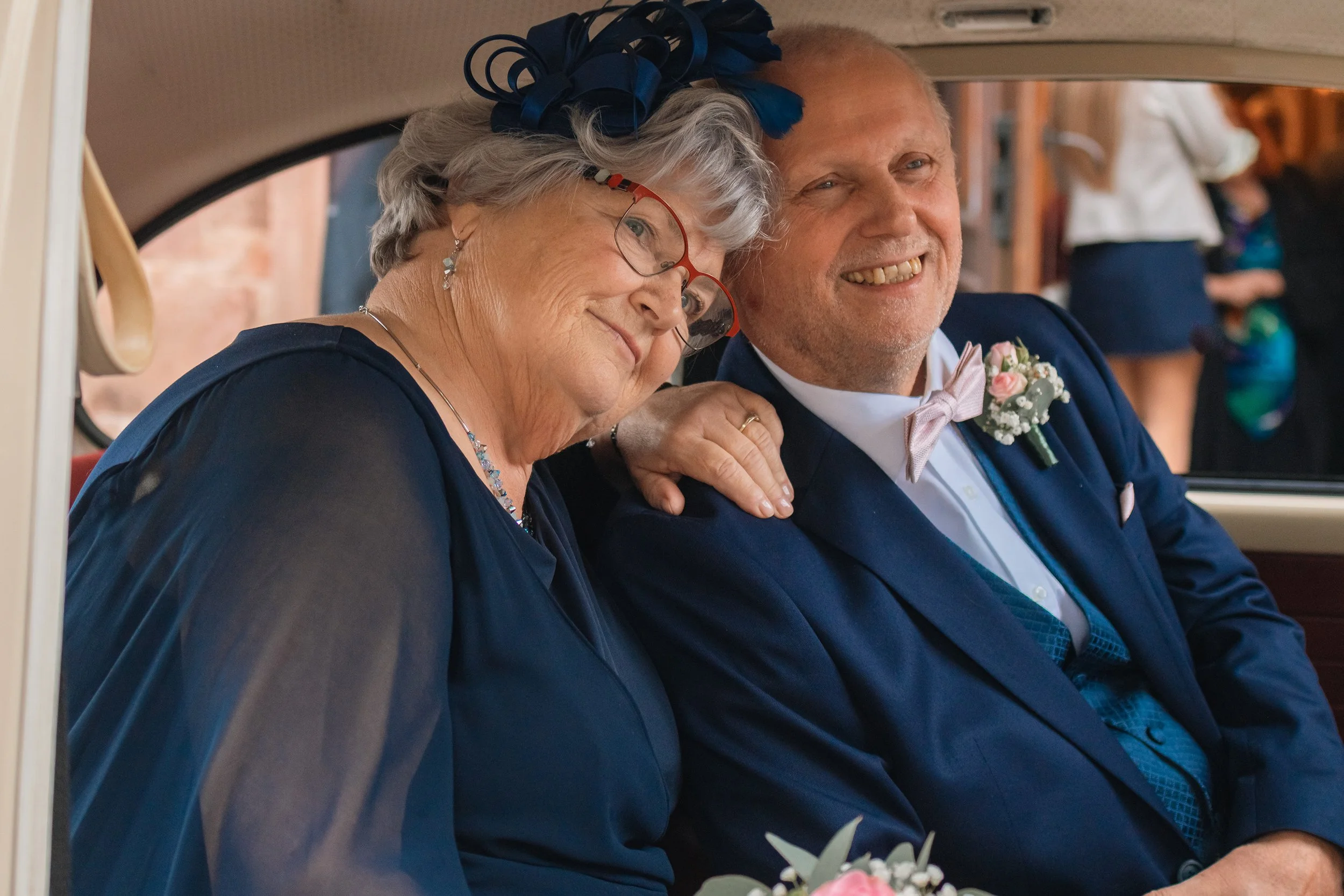 Ein älteres Paar in formeller Kleidung sitzt in einem Auto, lächelt und wirkt glücklich, wahrscheinlich an einem besonderen Anlass wie einer Hochzeit.