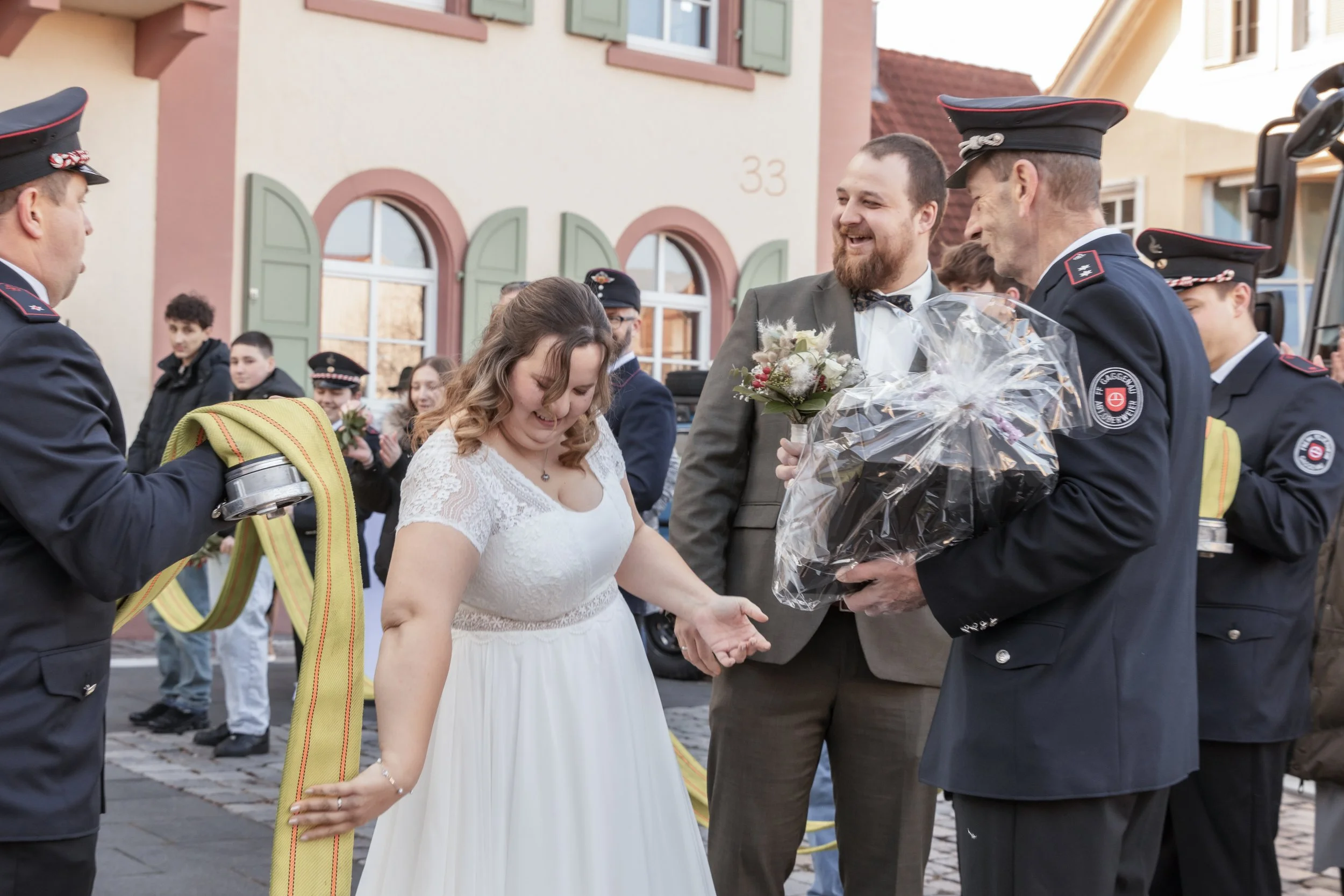 Hochzeitspaar umgeben von Feuerwehrleuten bei einer Feier im Freien. Die Braut trägt ein weißes Kleid, der Bräutigam einen grauen Anzug, und Feuerwehrleute übergeben Geschenke und Blumen.