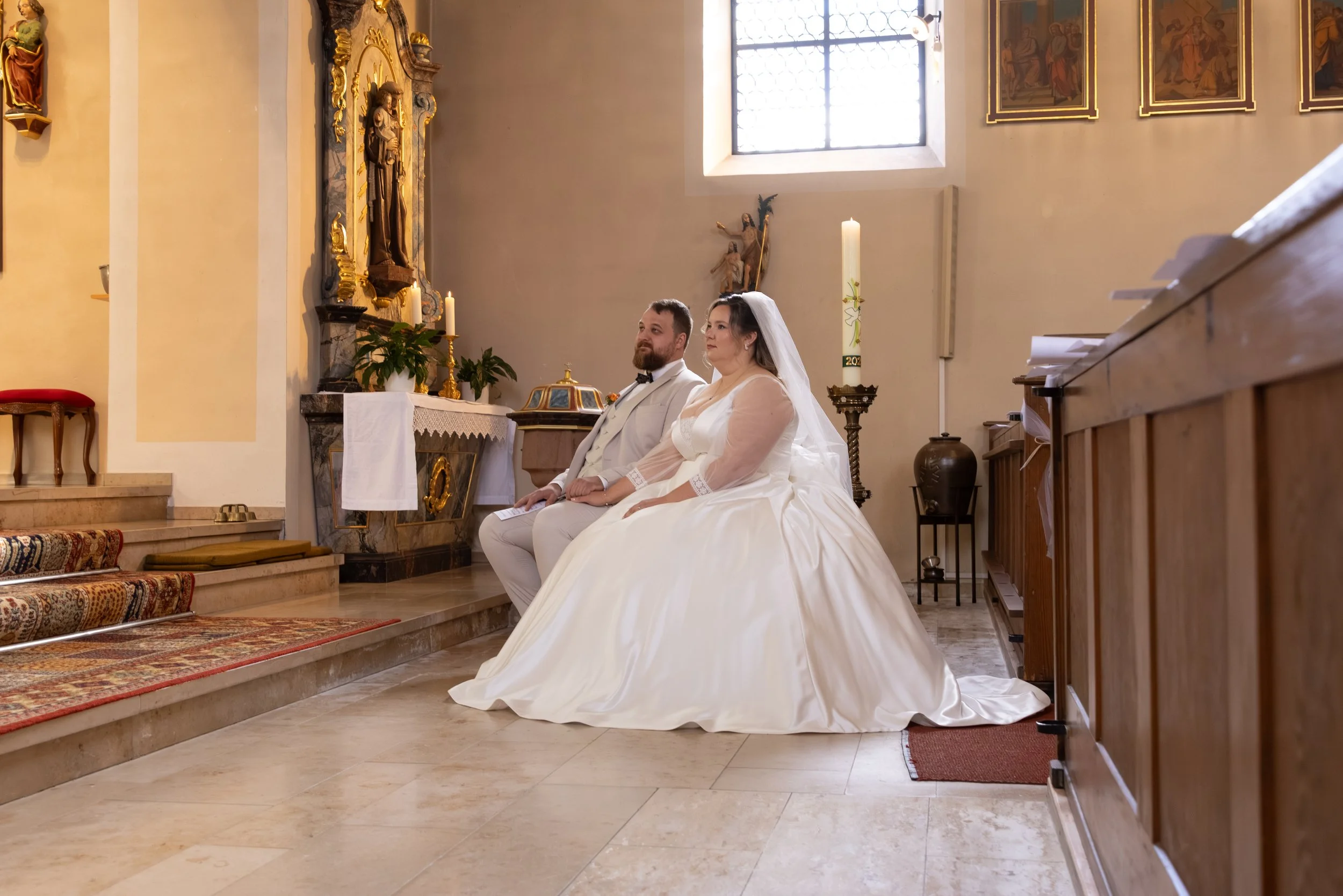 Ein Brautpaar sitzt in einer Kirche während einer Trauungszeremonie, die Braut trägt ein weißes Hochzeitskleid mit Schleier, der Bräutigam einen weißen Anzug.