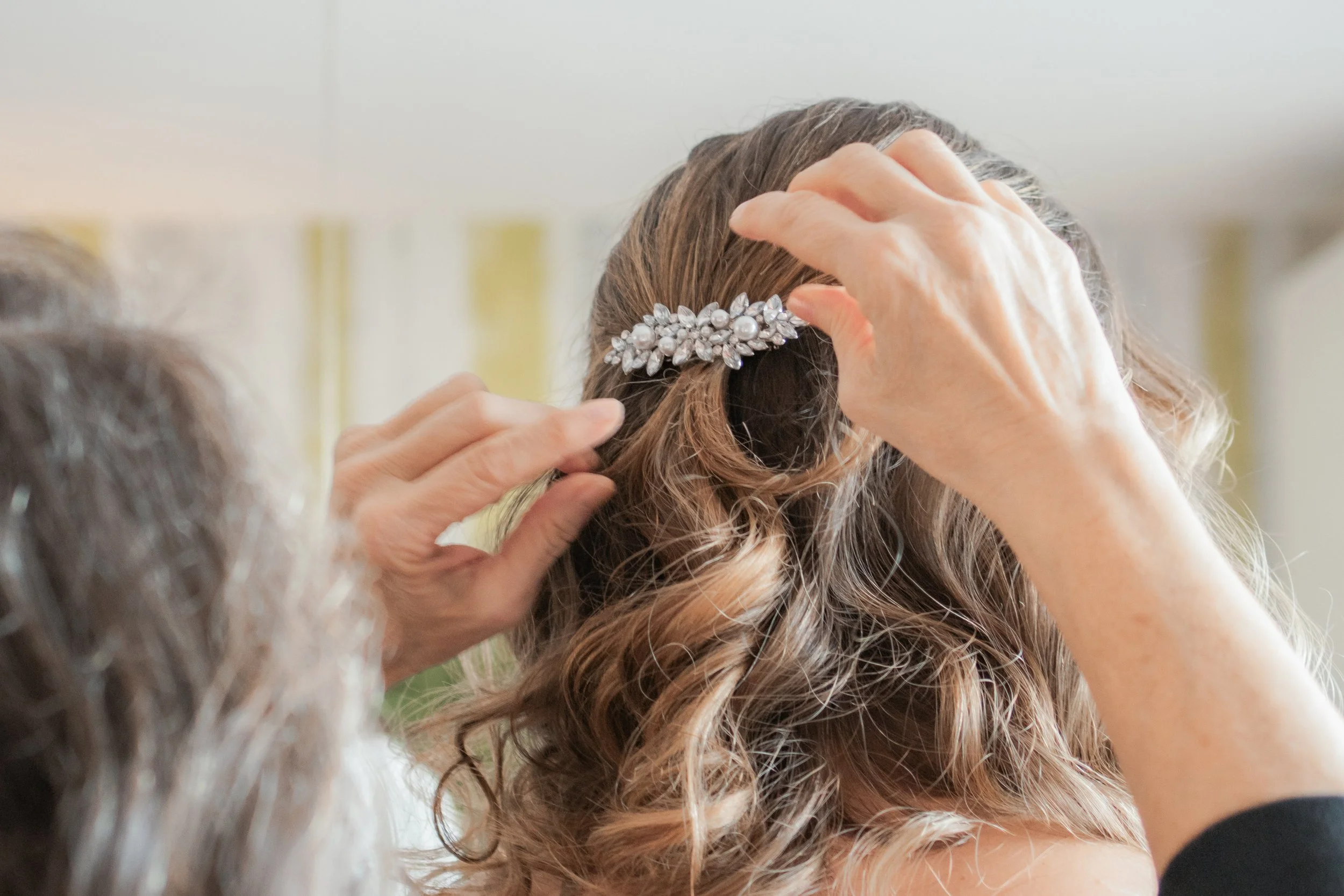 Eine Person steckt einer Frau einen eleganten Haarclip mit Perlen und Blumenmuster in die Locken ihres Haares.