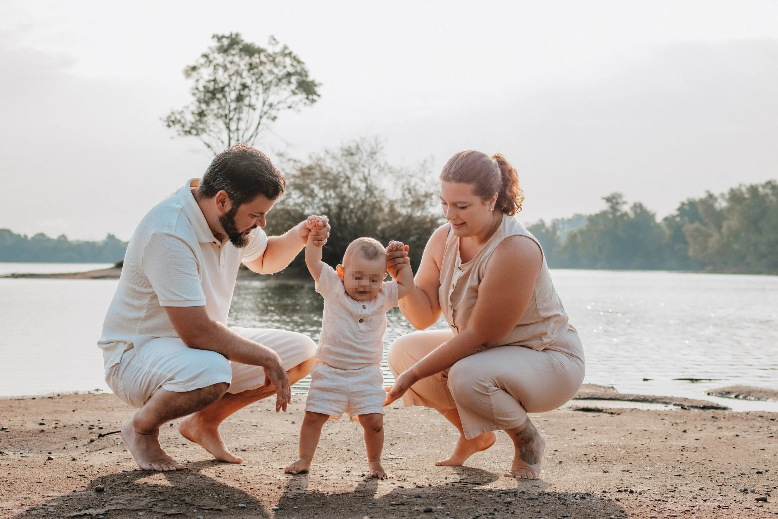 Ein glückliches Familienfoto von einem Mann und einer Frau, die ihrem kleinen Kind beim Gehen am Flussufer helfen, während sie lacht und die Natur im Hintergrund genießen.