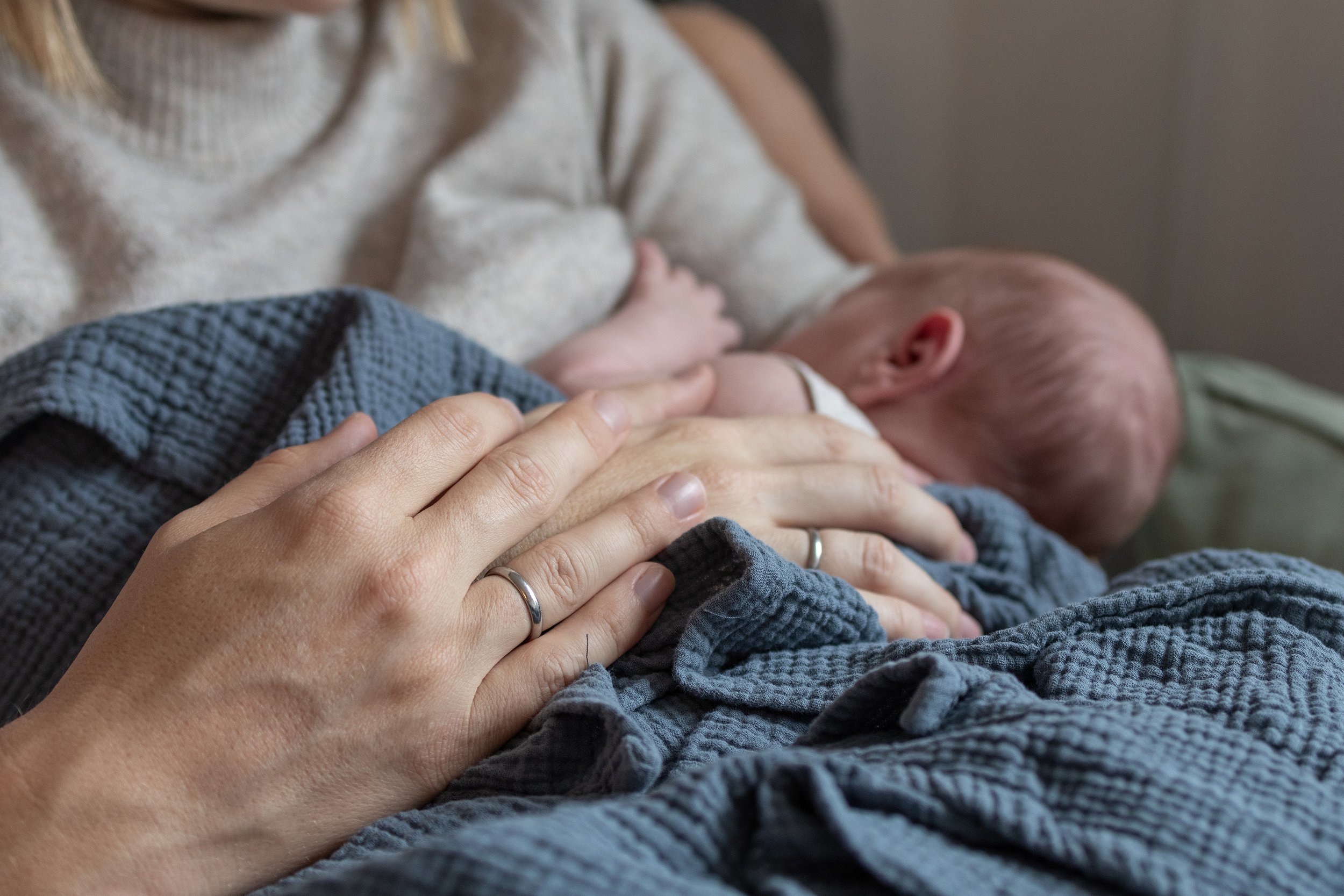Eine Person hält ein Baby, das an der Brust gestillt wird, auf einem Bett. Die Person trägt ein Silberring und hat die Hände auf das Baby gelegt.