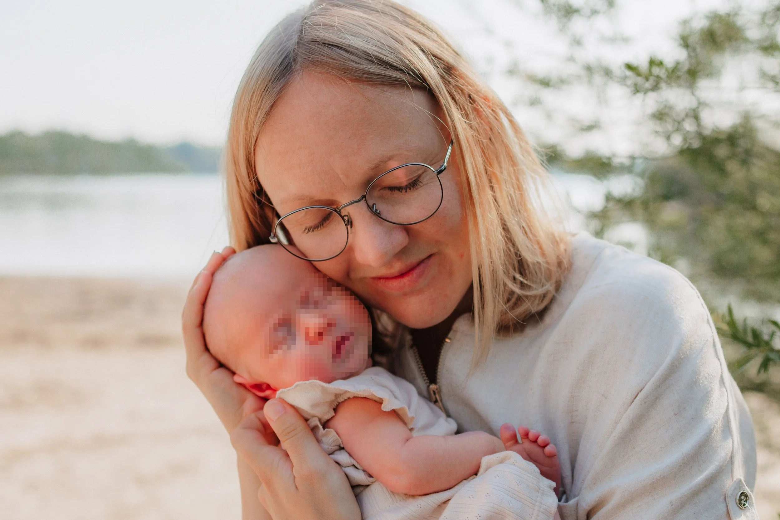 Eine Frau umarmt ein neugeborenes Baby und entspannt sich in der Natur in der Nähe eines Wassers. Die Frau trägt Brille und ein helles Jackett.