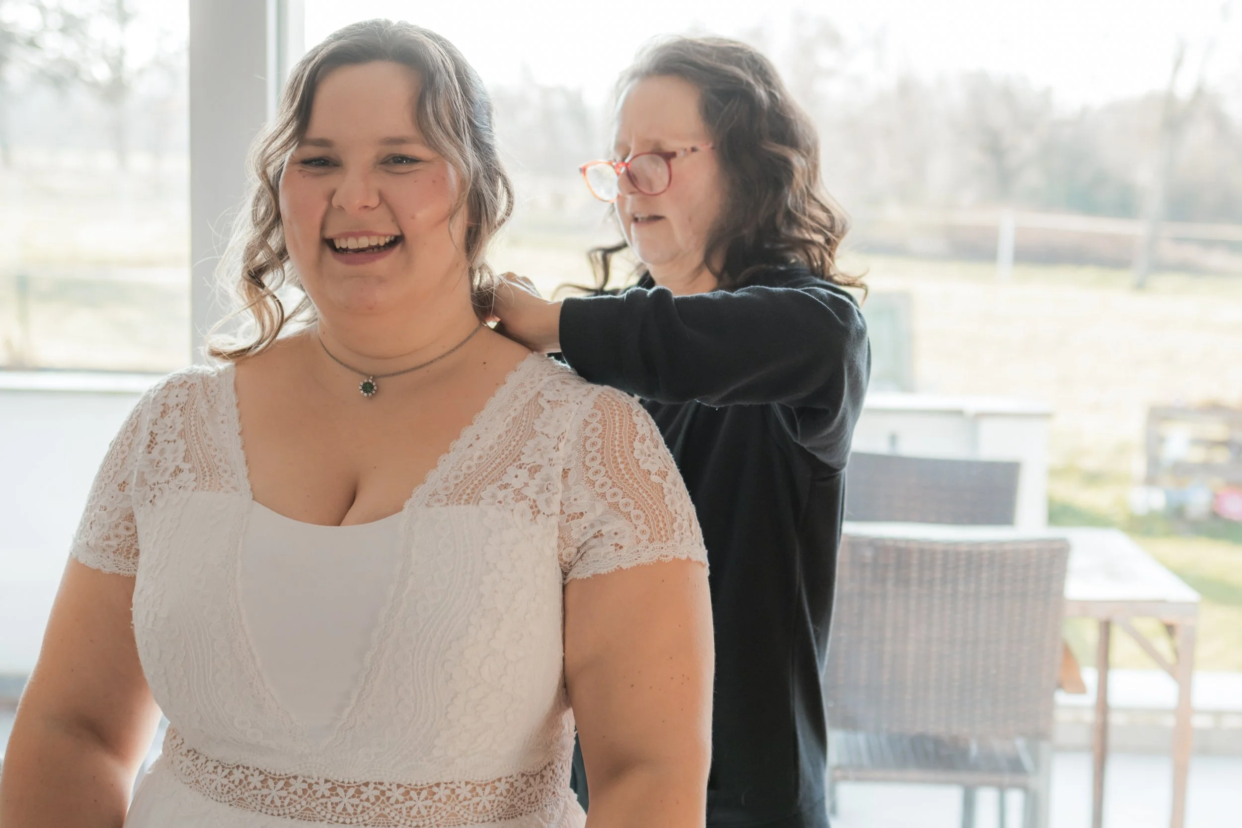 Eine junge Frau in einem weißen Spitzenkleid erlebt einen Moment der Freude, während eine ältere Frau mit Brille sie im Hintergrund anlächelt und ihr den Rücken massiert.
