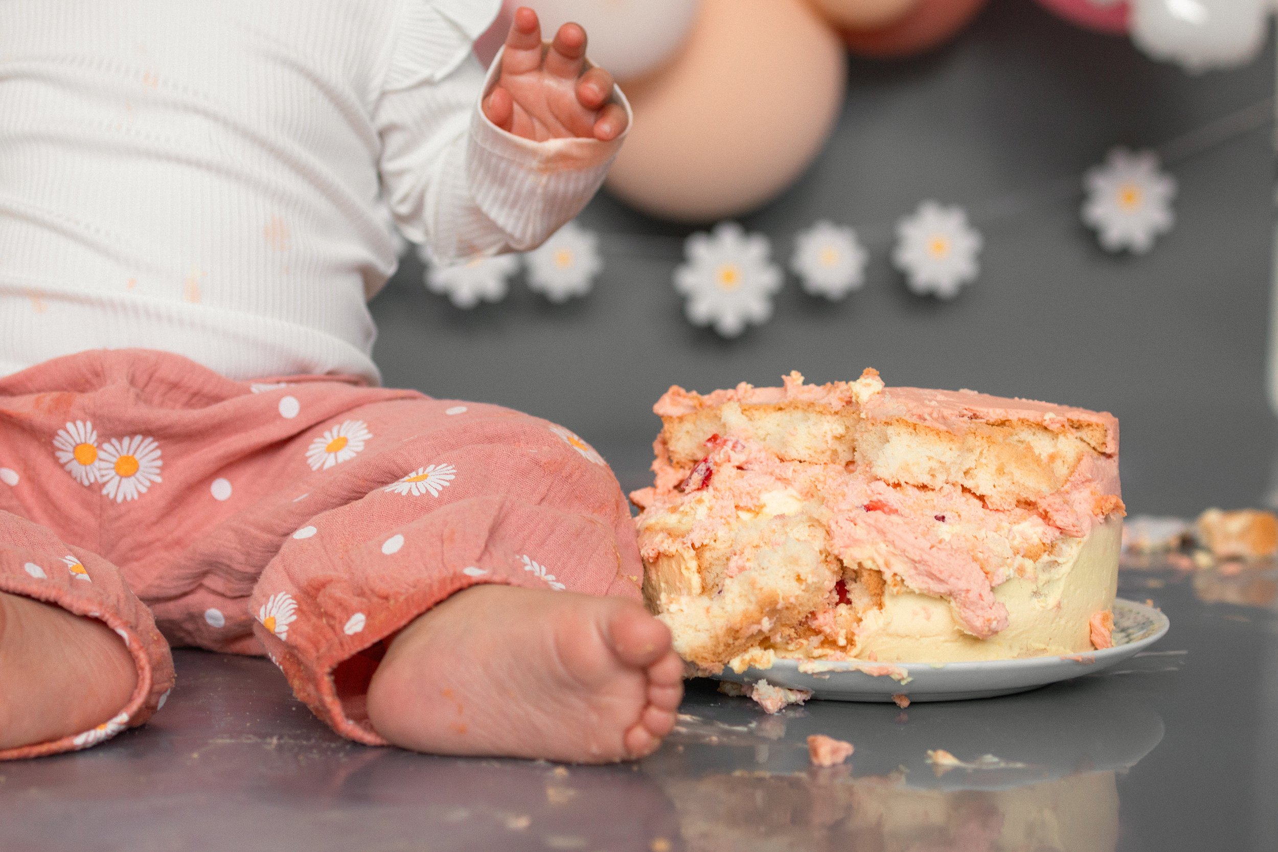 Ein Baby sitzt auf einem Tisch vor einem halbfertigen Kuchen mit rosa und weißem Frosting, umgeben von weißen Gänseblümchen-Dekorationen.