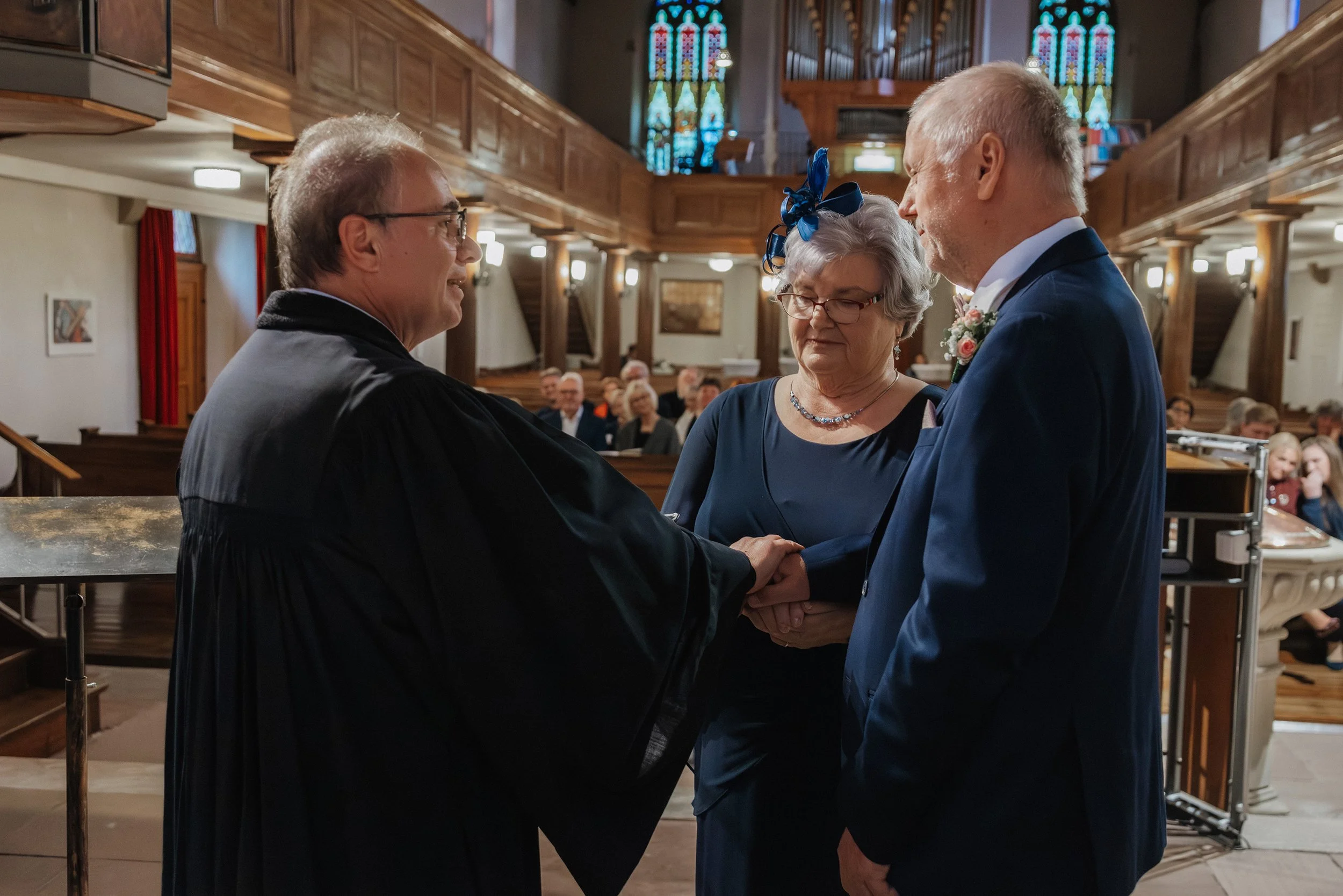 Ein älteres Paar bei einer Hochzeit steht vor einem Pastor in einer Kirche, während sie sich die Hände halten. Die Gäste sitzen im Hintergrund, die Kirche ist rustikal mit Holzdekorationen und bunten Kirchenfenstern.