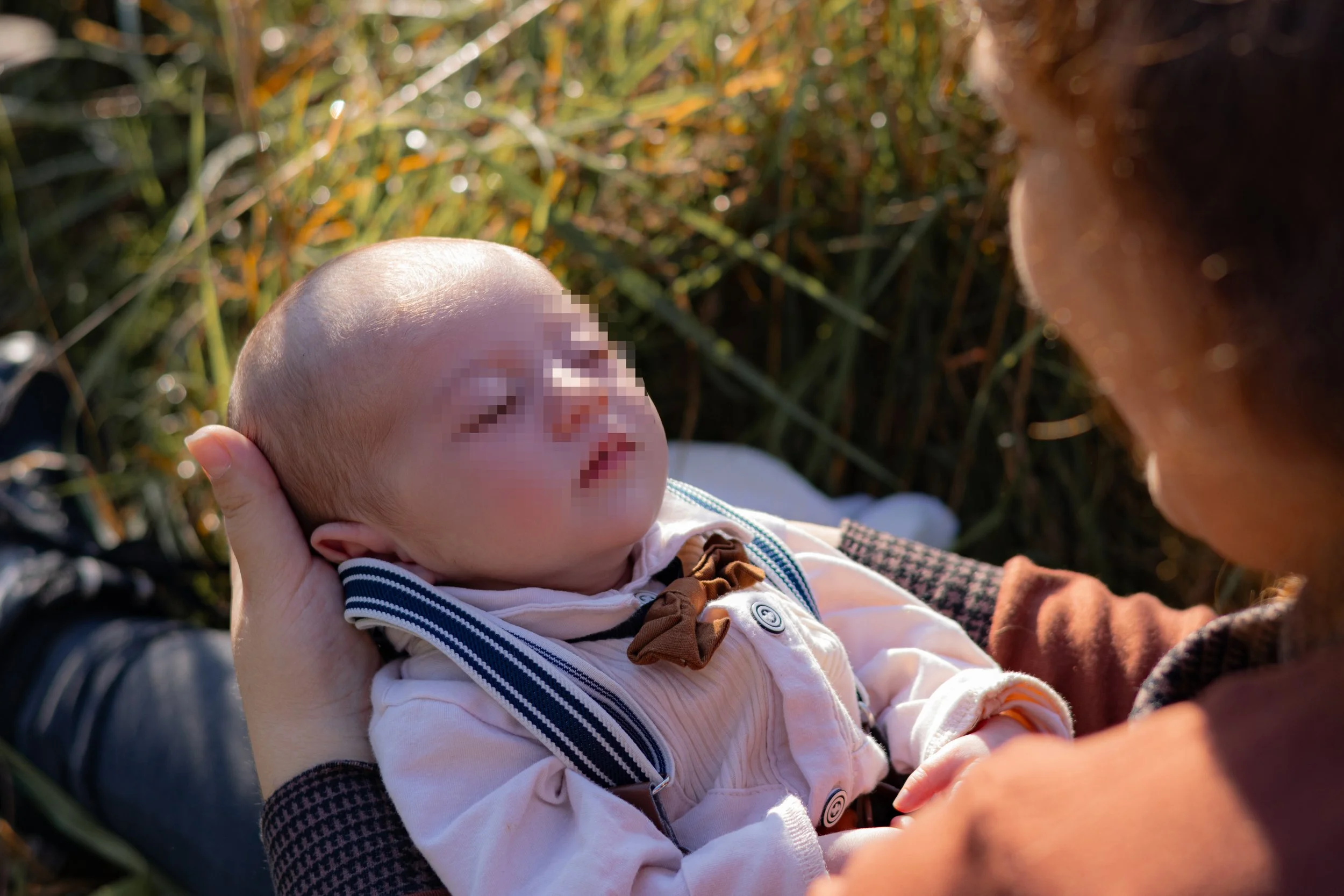 Ein Baby wird im Freien in den Armen einer Frau gehalten, im Hintergrund hohe Gräser, Sonnenlicht und Natur.