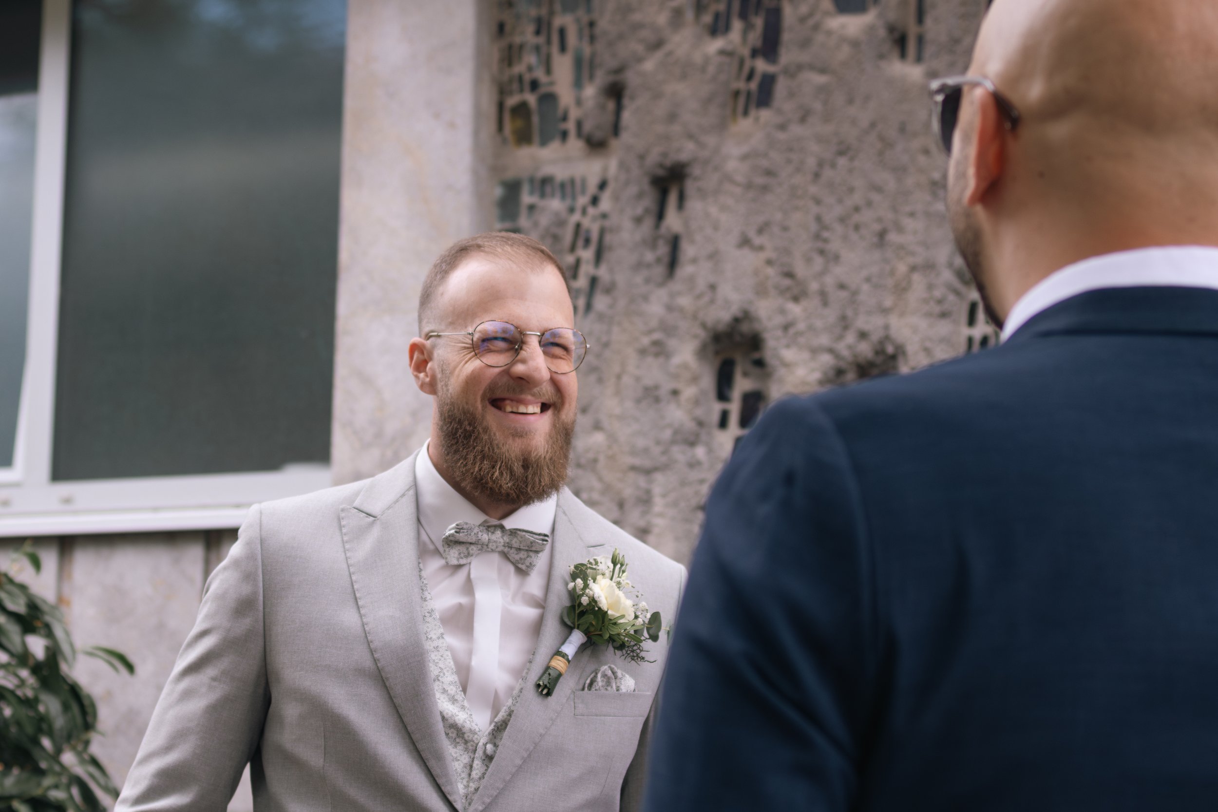 Zwei Männer bei einer Hochzeit, einer trägt einen hellgrauen Anzug mit Fliege und Brille, der andere trägt einen dunklen Anzug, beide lächeln und unterhalten sich draußen vor einer Steinmauer.