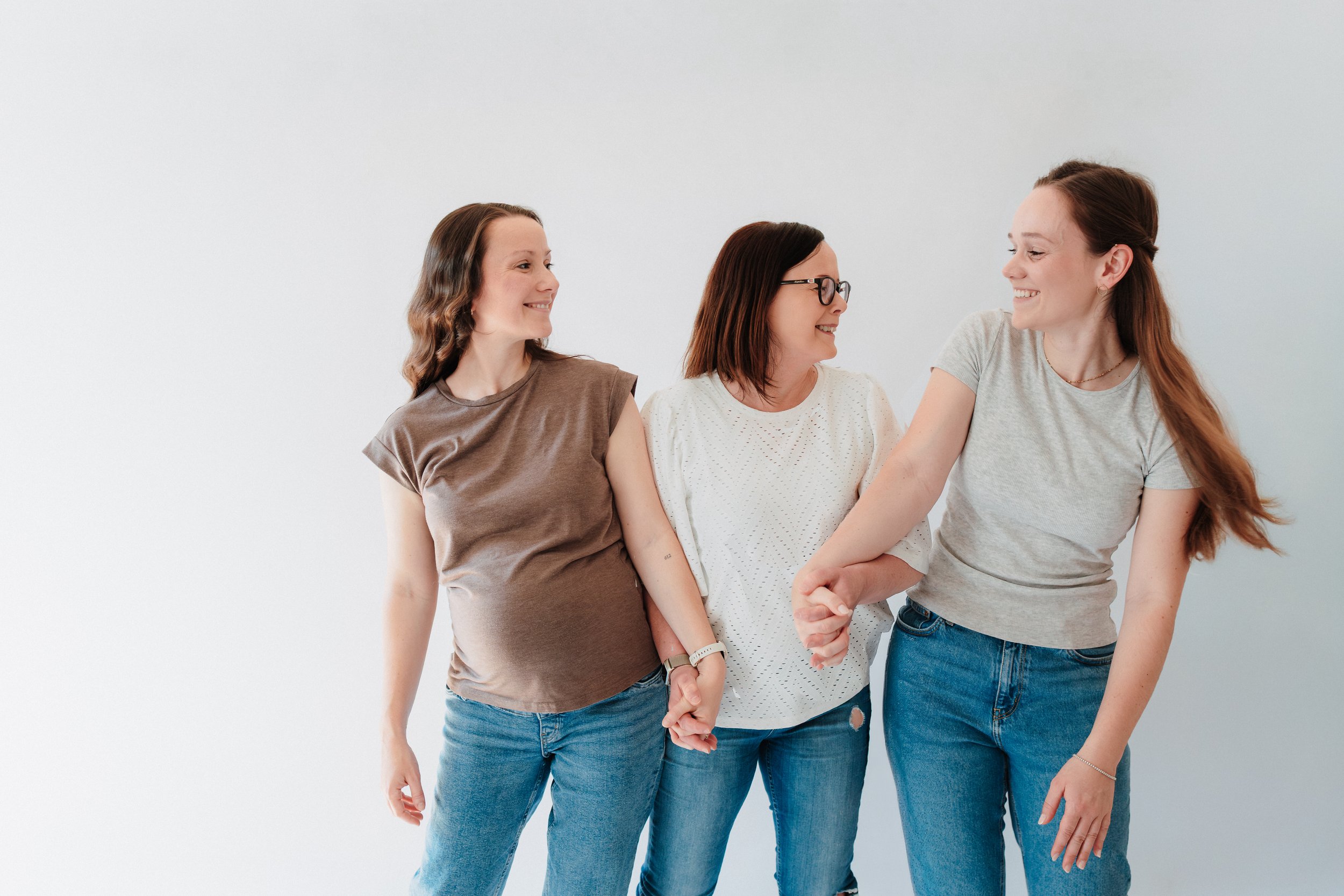 Drei Frauen stehen nebeneinander und halten sich an den Händen. Sie lachen und schauen sich an, vor einem weißen Hintergrund.