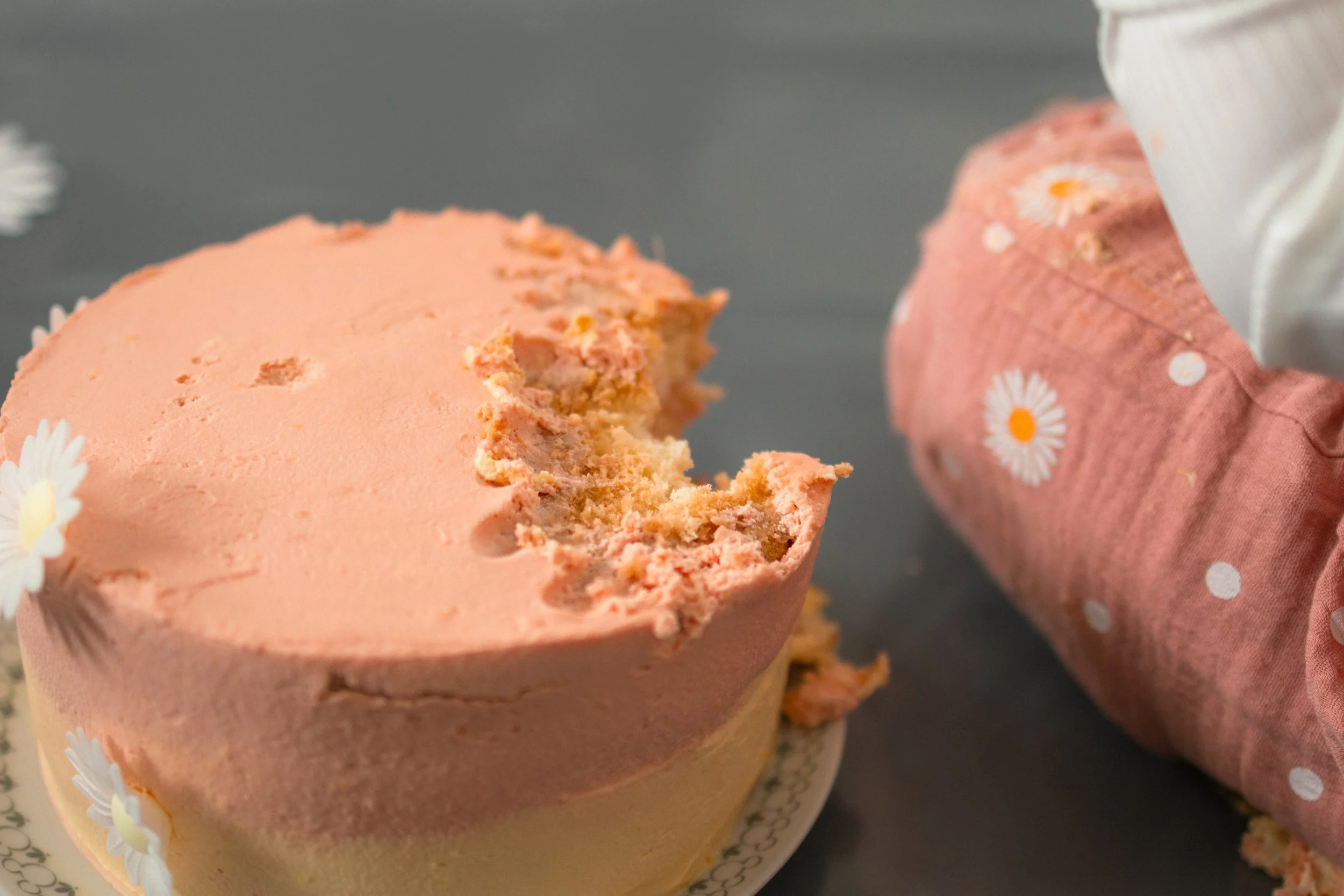 Ein halbfertiger Kuchen mit rosa Glasur und Dekorationen aus kleinen weißen Blumen, daneben ein in rosa mit weißen Punkten eingepackter Kuchen.