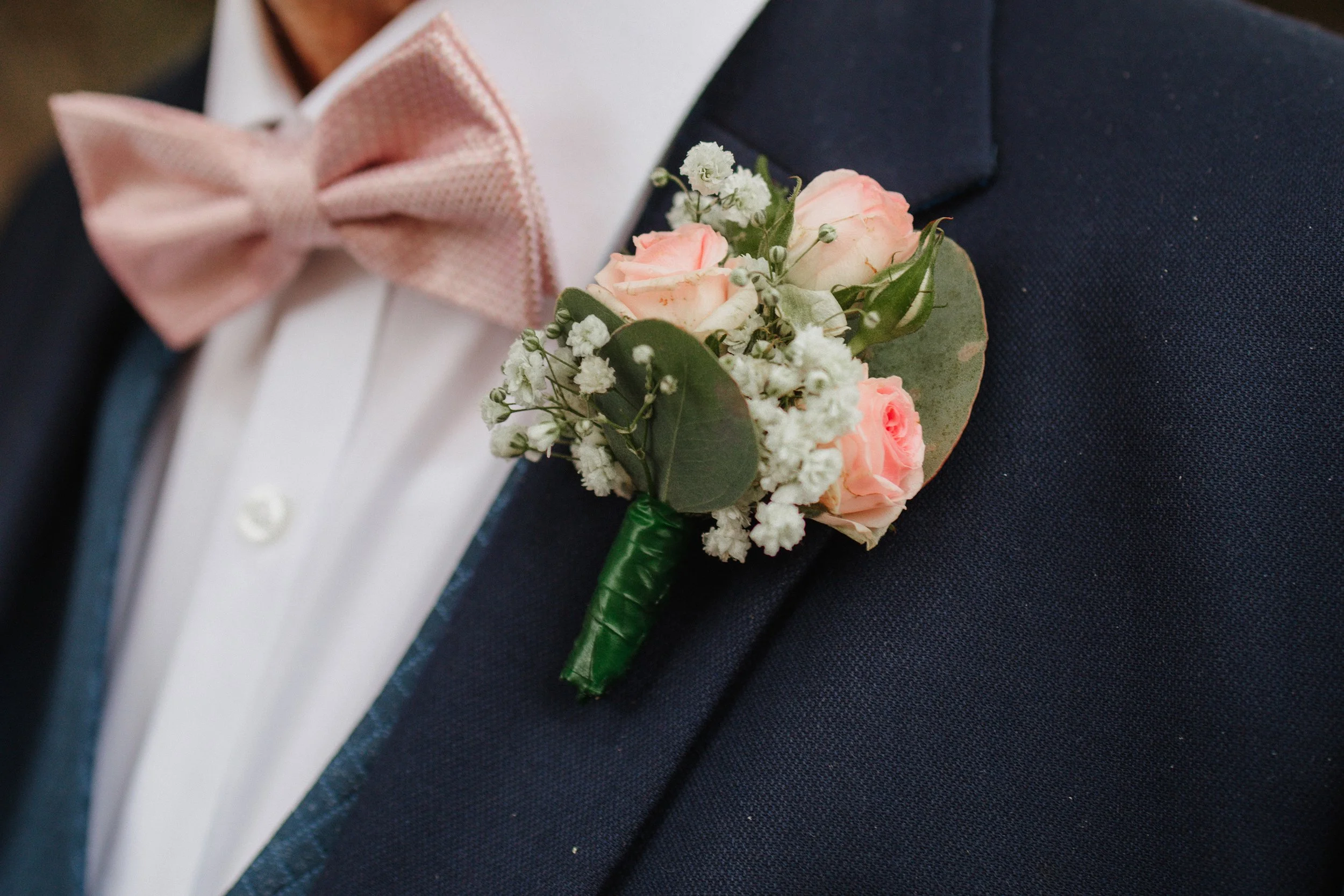 Nahaufnahme eines Ansteckblumens mit rosa Rosen, weißen Blumen und grünen Blättern, getragen an der Jacke eines Mannes mit weißem Hemd und rosa Fliege.
