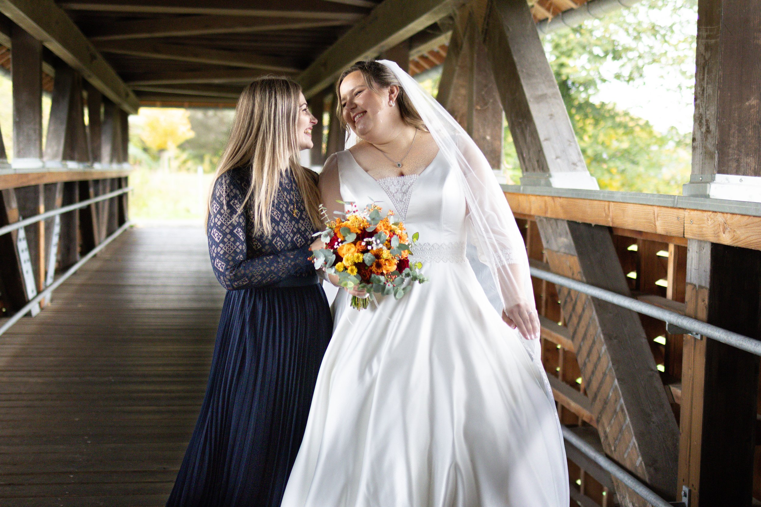 Zwei Frauen, eine in einem weißen Brautkleid mit Schleier und die andere in einem schwarzen Kleid, stehen sich gegenüber und lachen bei einer Hochzeitsfeier im Freien auf einer Holzbrücke.