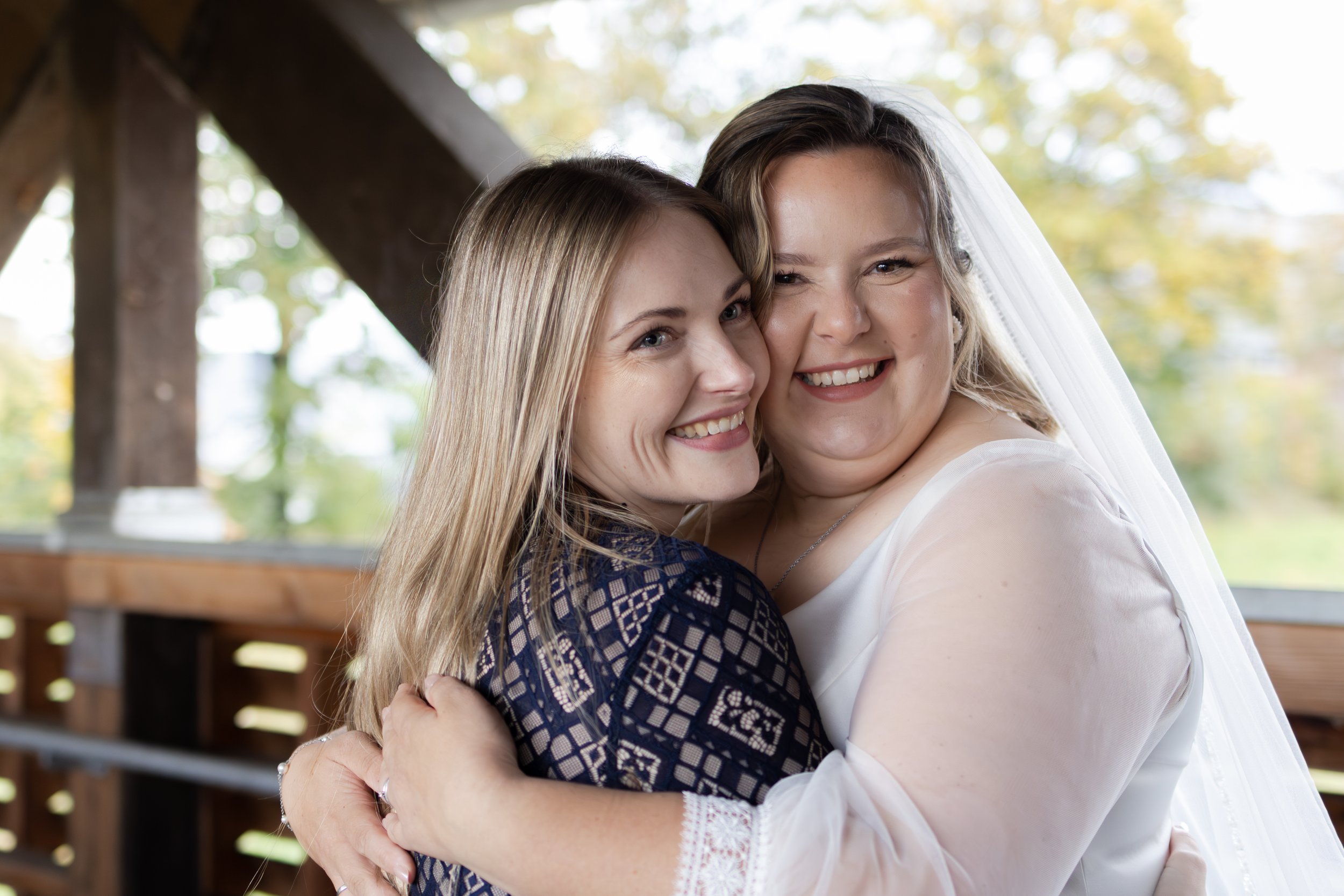Zwei Frauen umarmen sich bei einer Hochzeit im Freien, eine trägt ein Brautkleid mit Schleier, die andere ein dunkelblaues Kleid mit offenem Rücken.