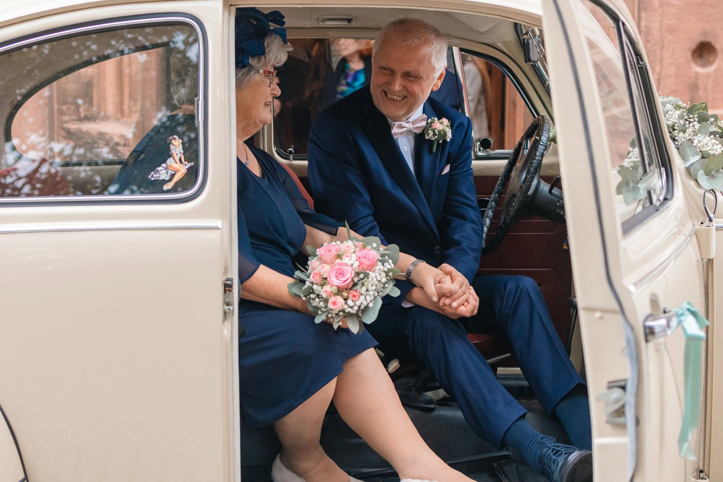 Ein älteres Paar, vermutlich Braut und Bräutigam, sitzen in einem Vintage-Auto, der Bräutigam hält die Hand der Braut. Beide tragen elegante Kleidung und lächeln glücklich. Die Braut hält einen Blumenstrauß aus rosa und weißen Blumen.