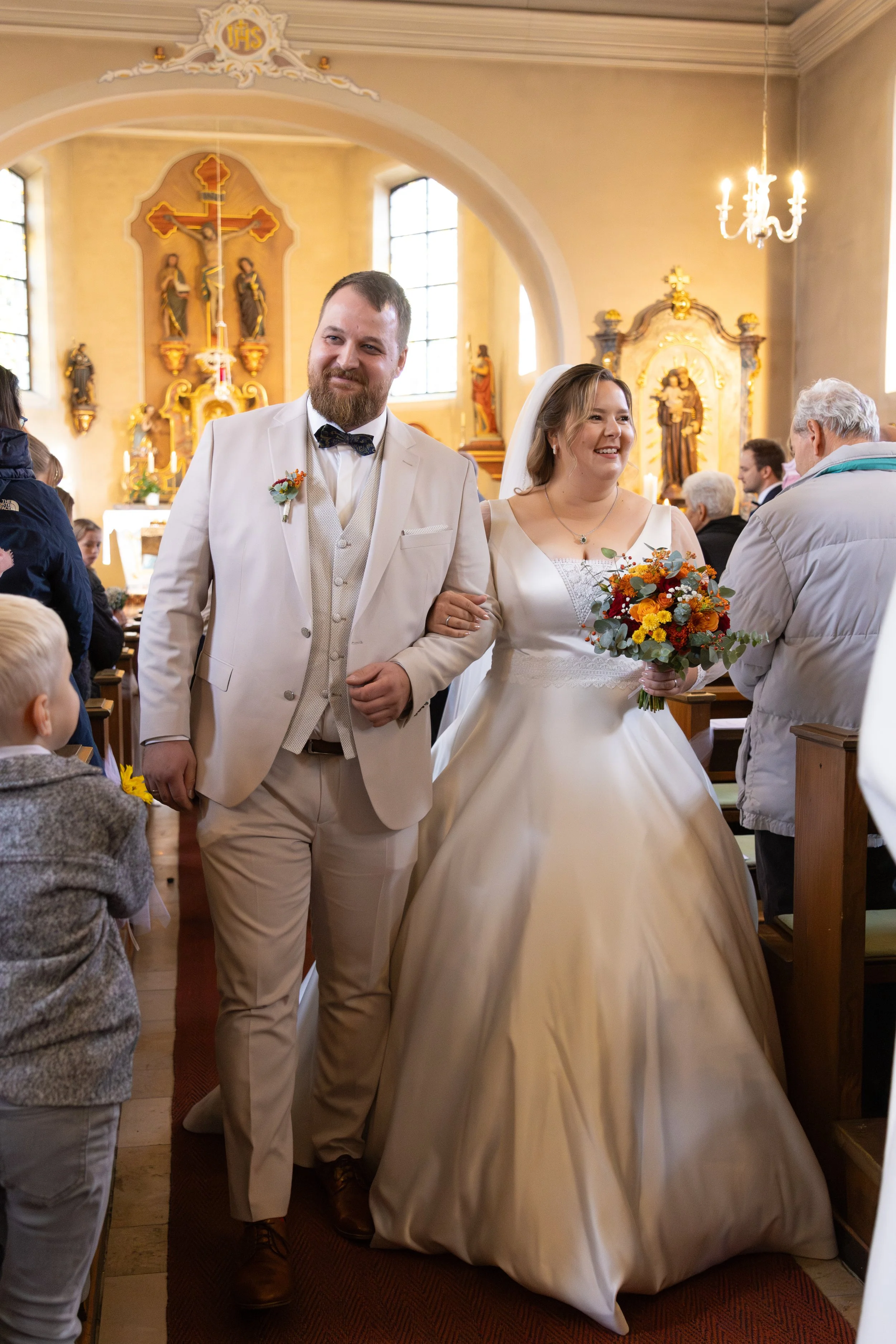 Braut und Bräutigam gehen in einer Kirche während ihrer Hochzeitszeremonie. Die Braut trägt ein weißes Hochzeitskleid und hält einen bunten Blumenstrauß, der Bräutigam trägt einen weißen Anzug mit Fliege. Im Hintergrund sind Hochzeitsgäste und religi