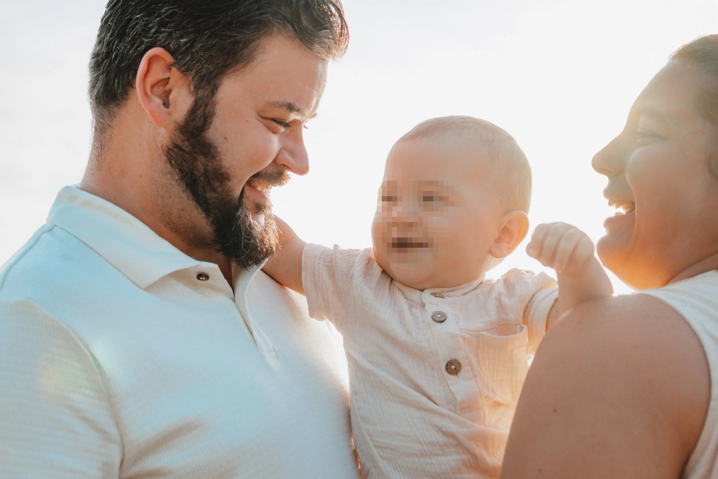 Ein glückliches Familienfoto von einem Mann, einer Frau und einem Baby, die draußen bei Sonnenuntergang lachen und Freude zeigen.