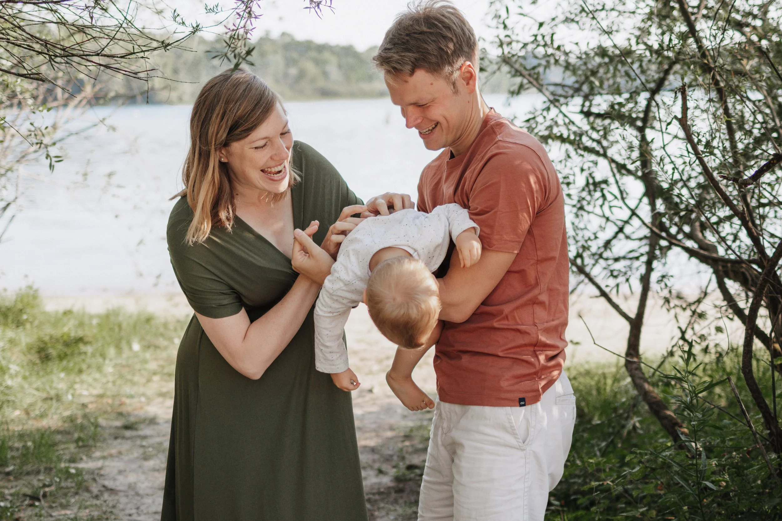 Ein Mann und eine Frau lachen und spielen mit einem Baby am See, umgeben von Bäumen.