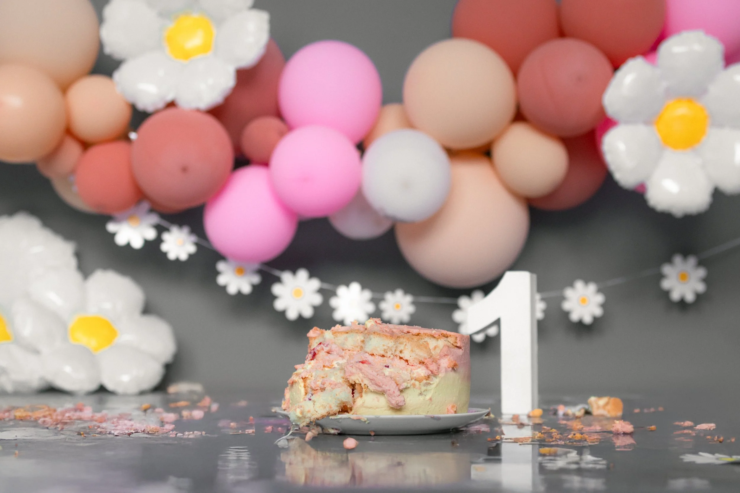 Ein halb gegessener Geburtstagstorte mit rosa und weißer Verzierung, umgeben von Dekorationen mit großen Ballon-Kugeln in Pastellfarben und Papierblumen. Ein weißes Zahl-1 steht neben der Torte.