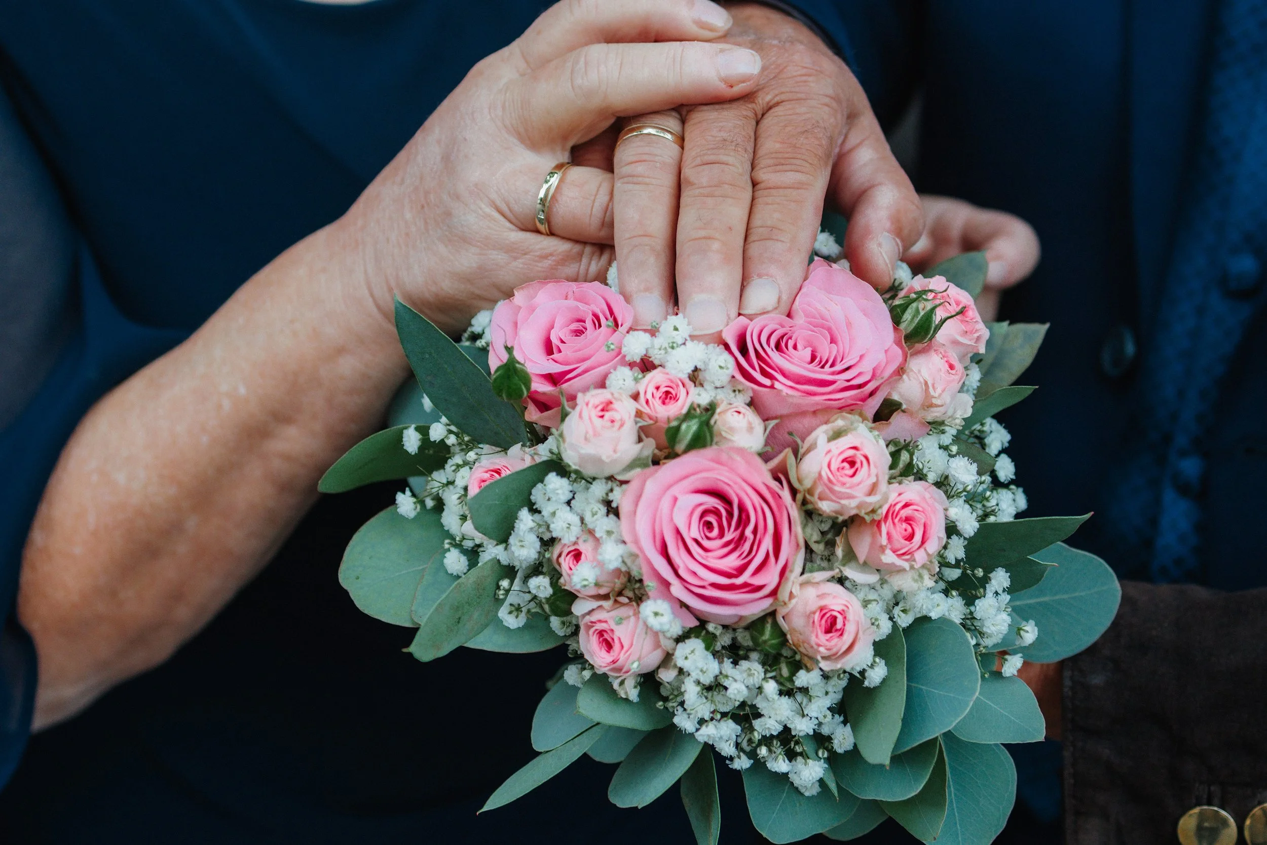 Näheaufnahme von Händen, die eine Brautstrauß halten, bestehend aus pinken Rosen, kleinen weißen Blumen und grünen Blättern.