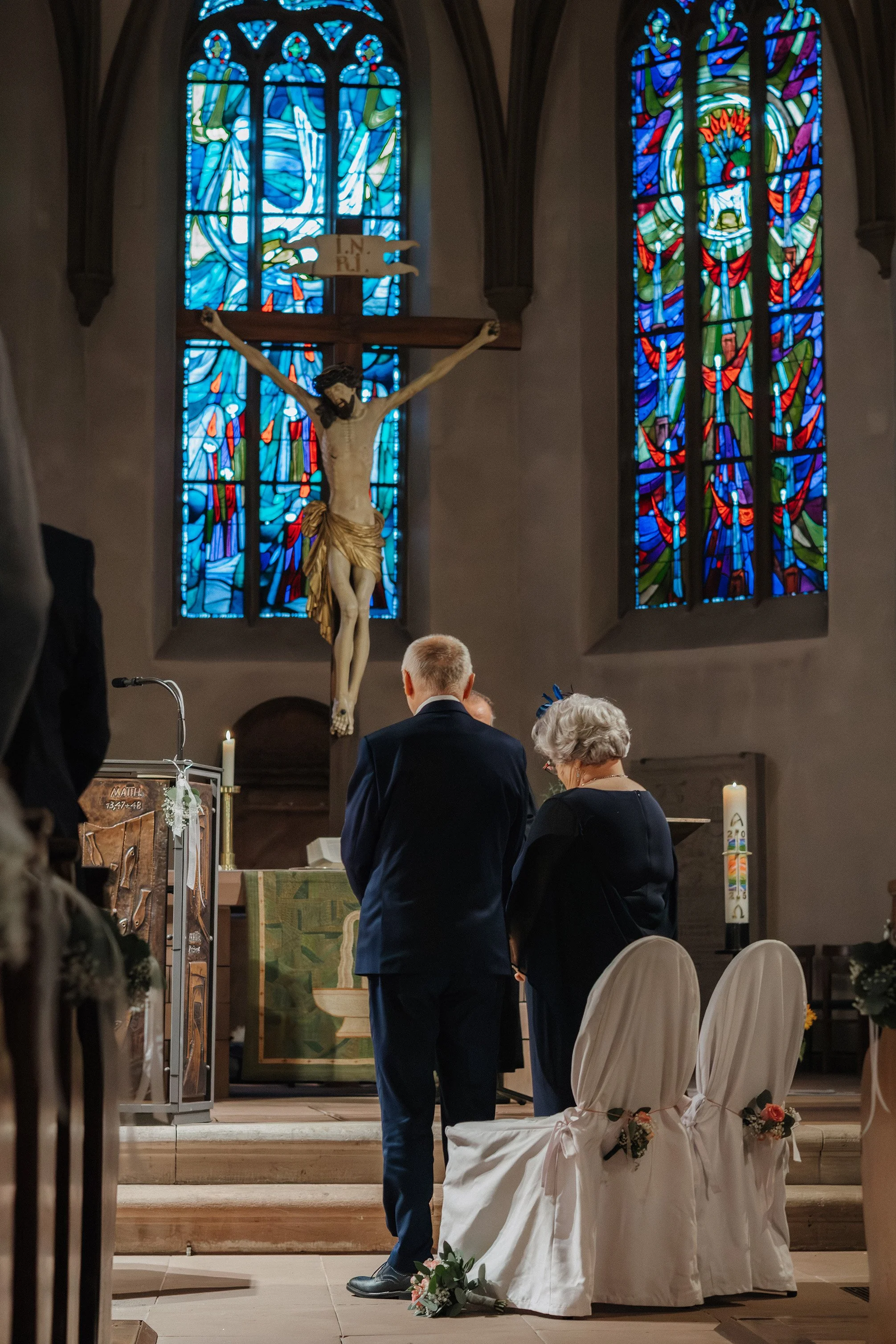 Zwei Menschen stehen in einer Kirche vor dem Altar, im Hintergrund ein Kreuz mit Jesus am Kreuz und bunte Kirchenfenster.