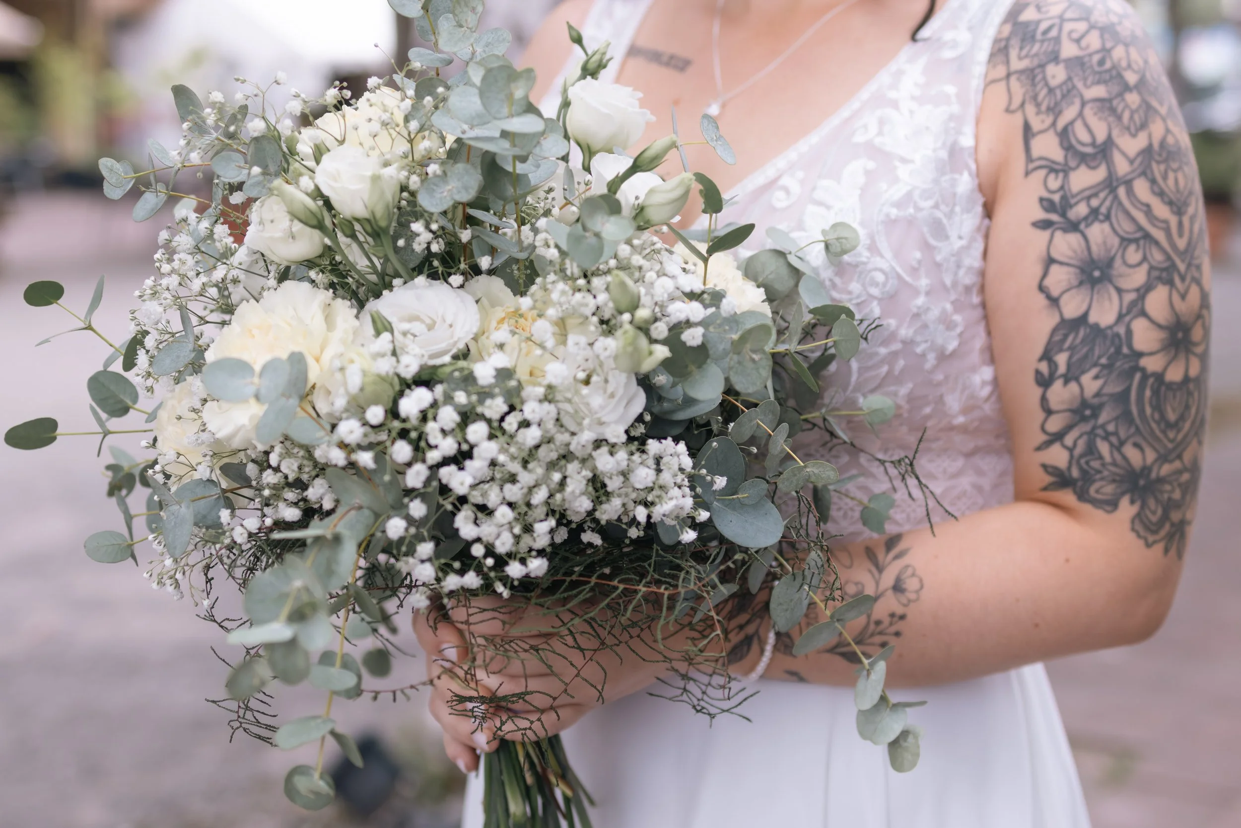 Eine Braut hält einen Brautstrauß aus weißen Blumen und grünen Blättern. Sie trägt ein weißes Hochzeitskleid mit Spitze und hat einen Tattoo-Arm.