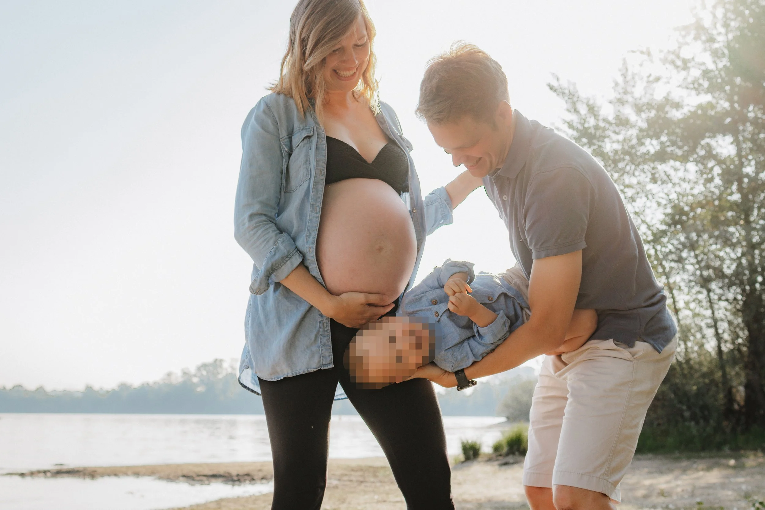 Familie steht am Flussufer, Mutter schwanger, Vater hält Baby mit pixelisiertem Gesicht, alle lachen gemeinsam.