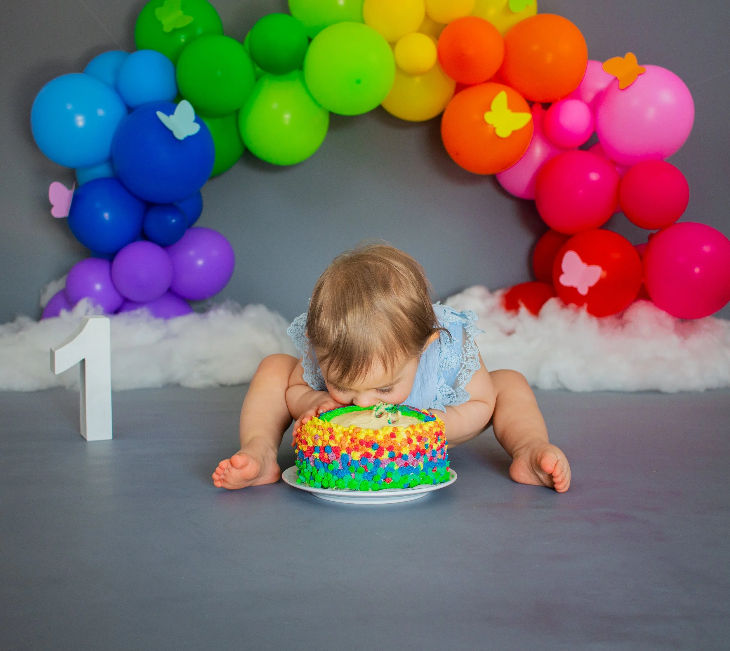 Ein Baby isst eine bunte Geburtstagstorte in einem Geburtstagsraum mit Ballons und einer Zahl 1 am Boden.