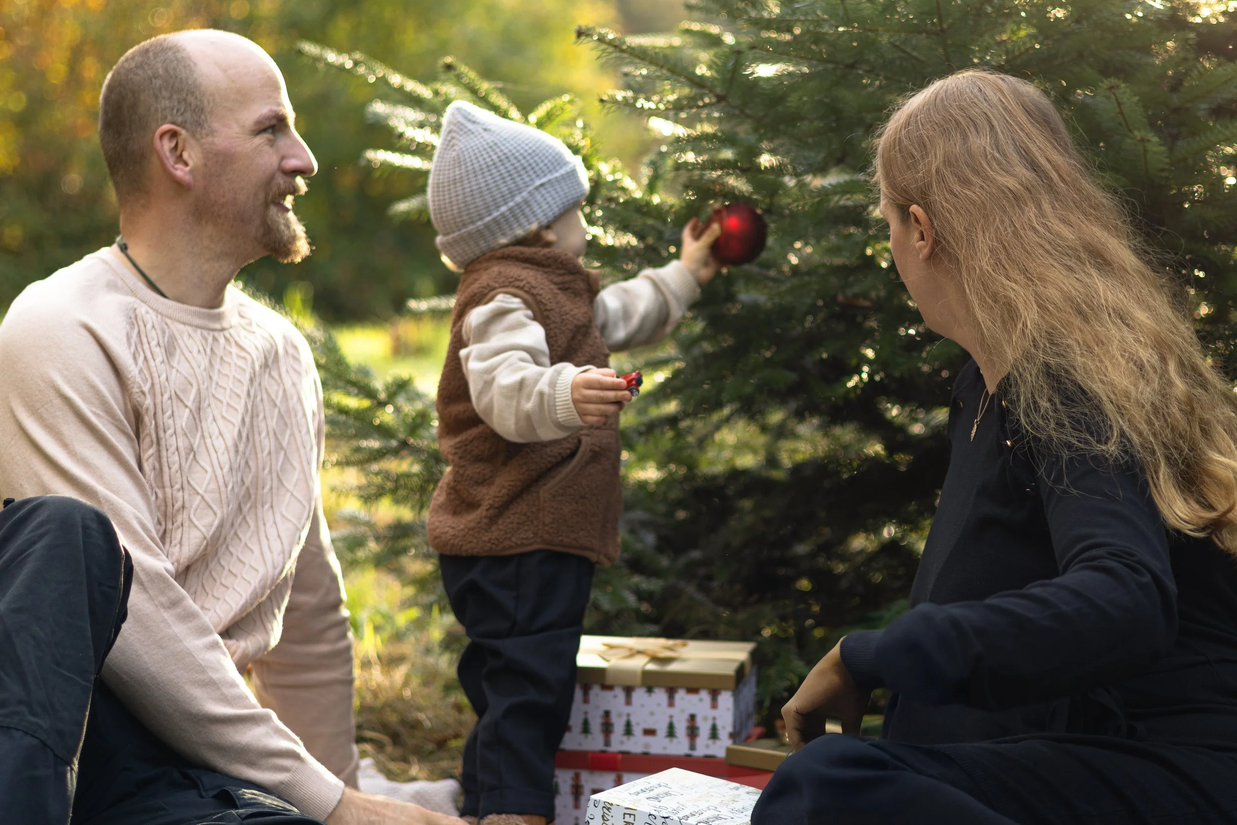 Familie beim Weihnachtsbaum schmücken im Freien, mit Geschenken im Hintergrund.