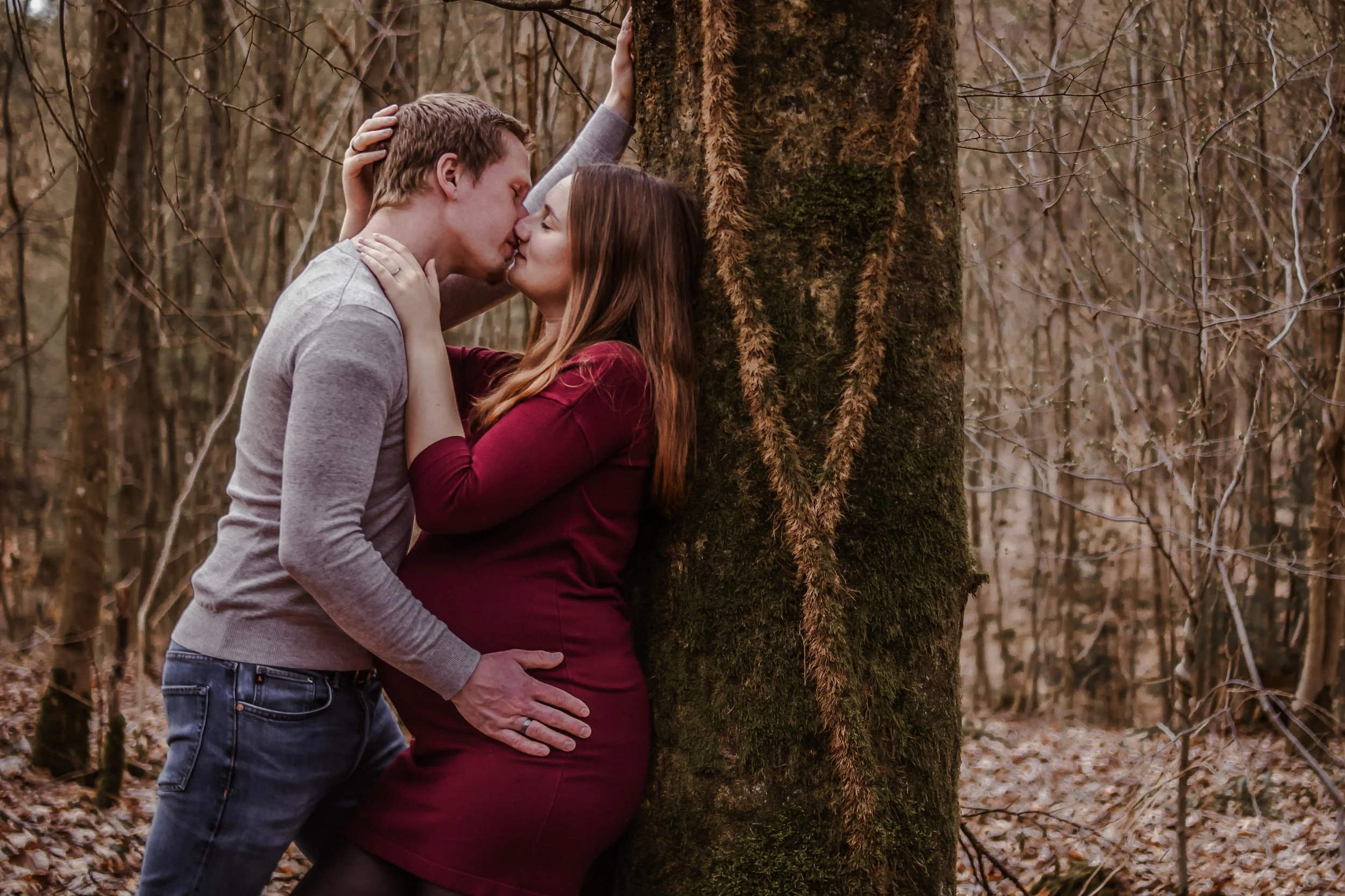 Ein Paar küsst sich in einem Wald, die Frau ist schwanger und trägt ein rotes Kleid, der Mann trägt ein graues Hemd.
