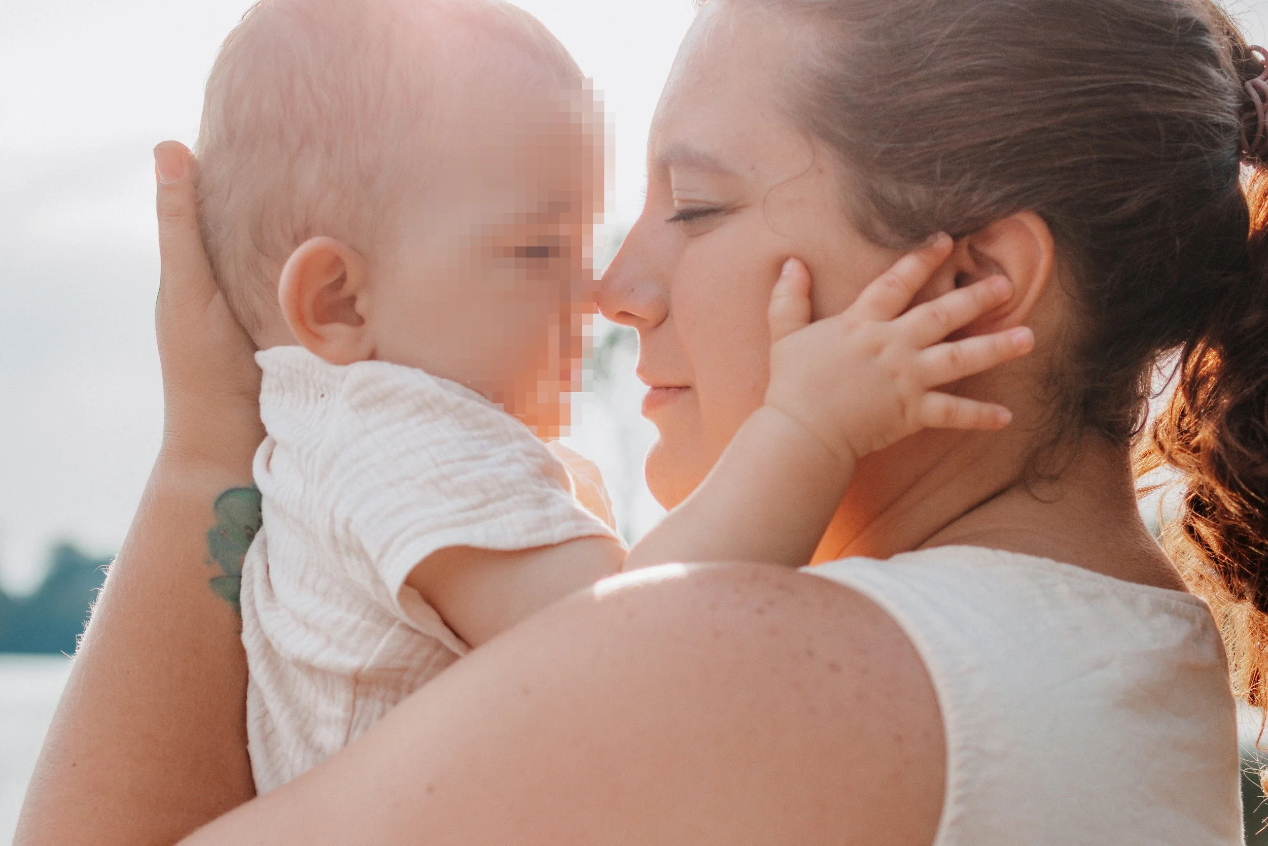 Eine Frau hält ein Baby nahe an ihr Gesicht, beide berühren sich mit den Nasen, die Augen sind geschlossen, im Hintergrund ist der Himmel sichtbar