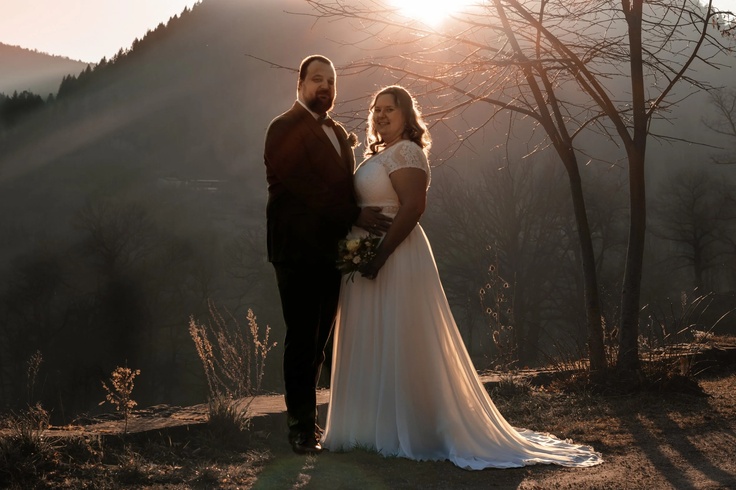 Ein Paar in Hochzeitskleidung steht im Sonnenuntergang, umgeben von Bäumen und einer Berglandschaft.