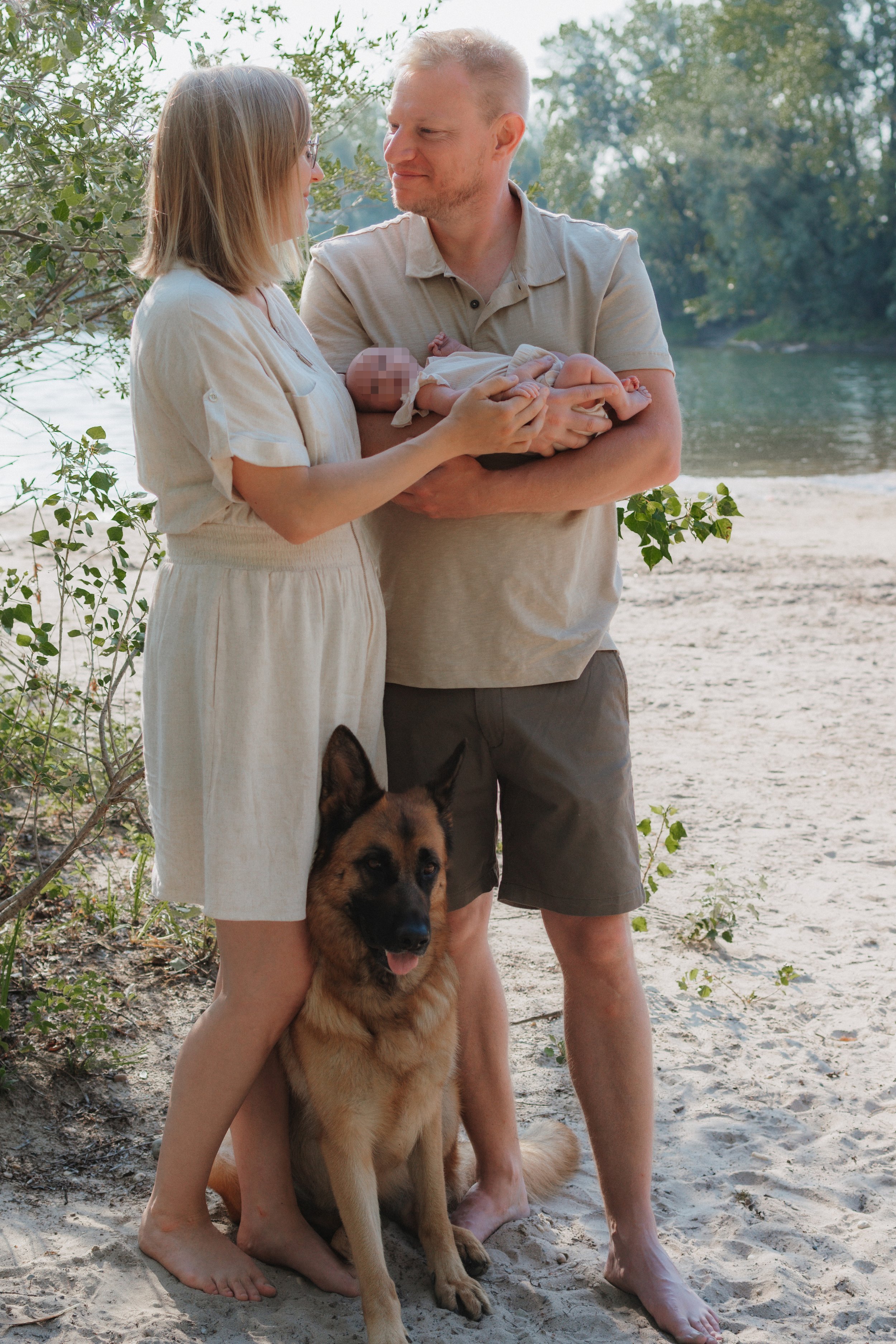 Ein Paar hält ein Baby am Flussufer, mit einem Deutschen Schäferhund sitzend vor ihnen, während sie sich liebevoll anschauen, bei Tageslicht im Freien.