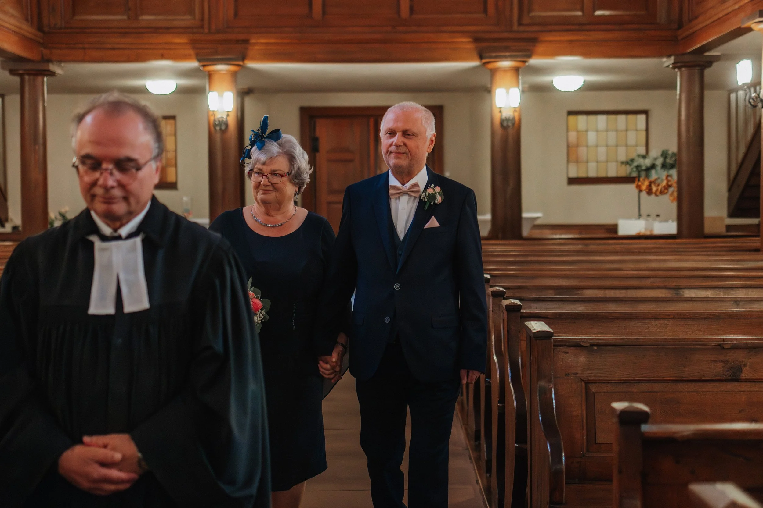 Ein Mann in einem schwarzen Anzug mit einer rosa Fliege, eine ältere Frau in einem dunklen Kleid mit einer blauen Schleife im Haar, und ein Mann in einem schwarzen Robe bei einer Hochzeitszeremonie in einer Kirche.