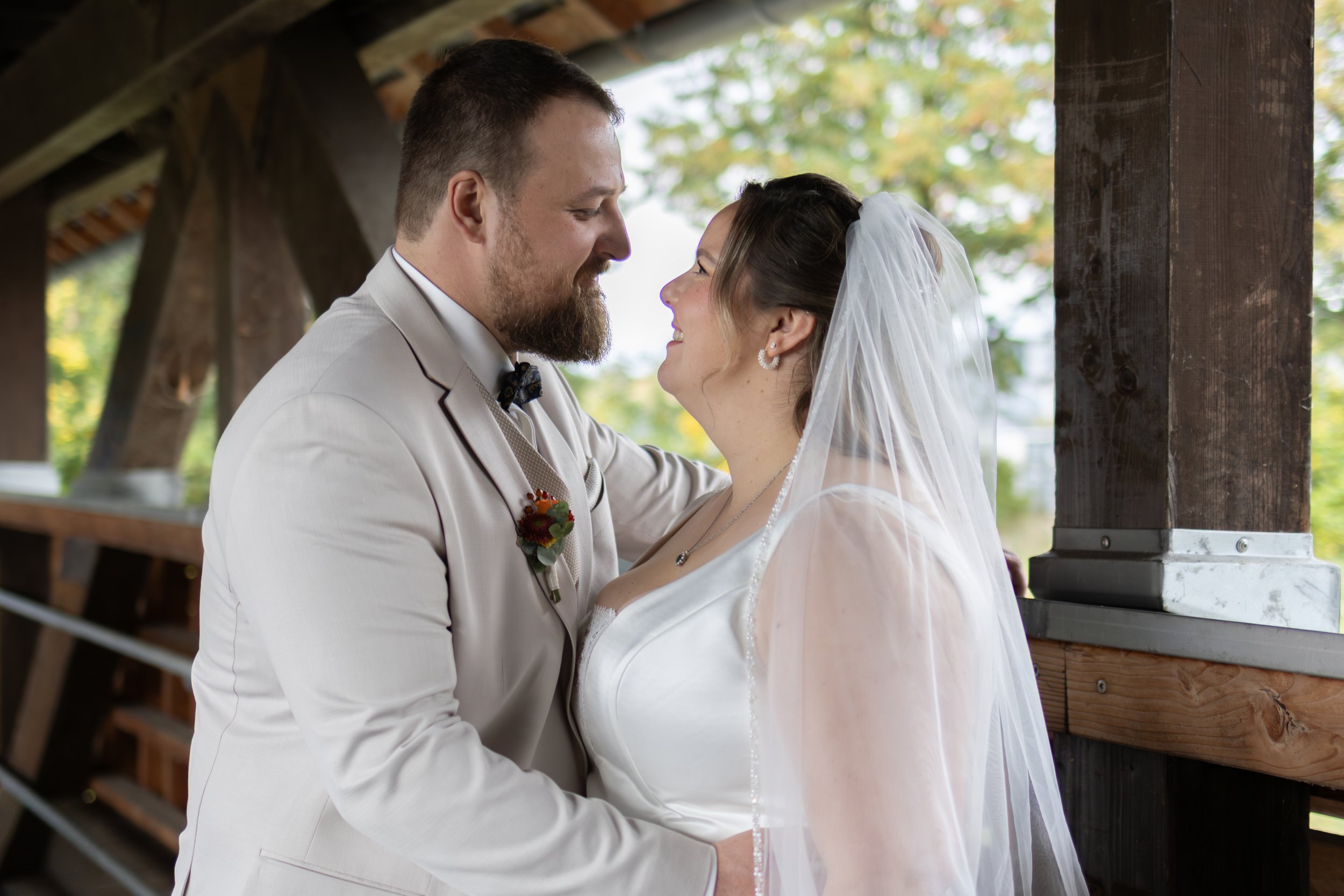 Ein Brautpaar steht sich bei einer Hochzeit gegenüber, lächelt sich an und hält sich an den Händen, im Hintergrund sind Holzbalken und Bäume sichtbar.