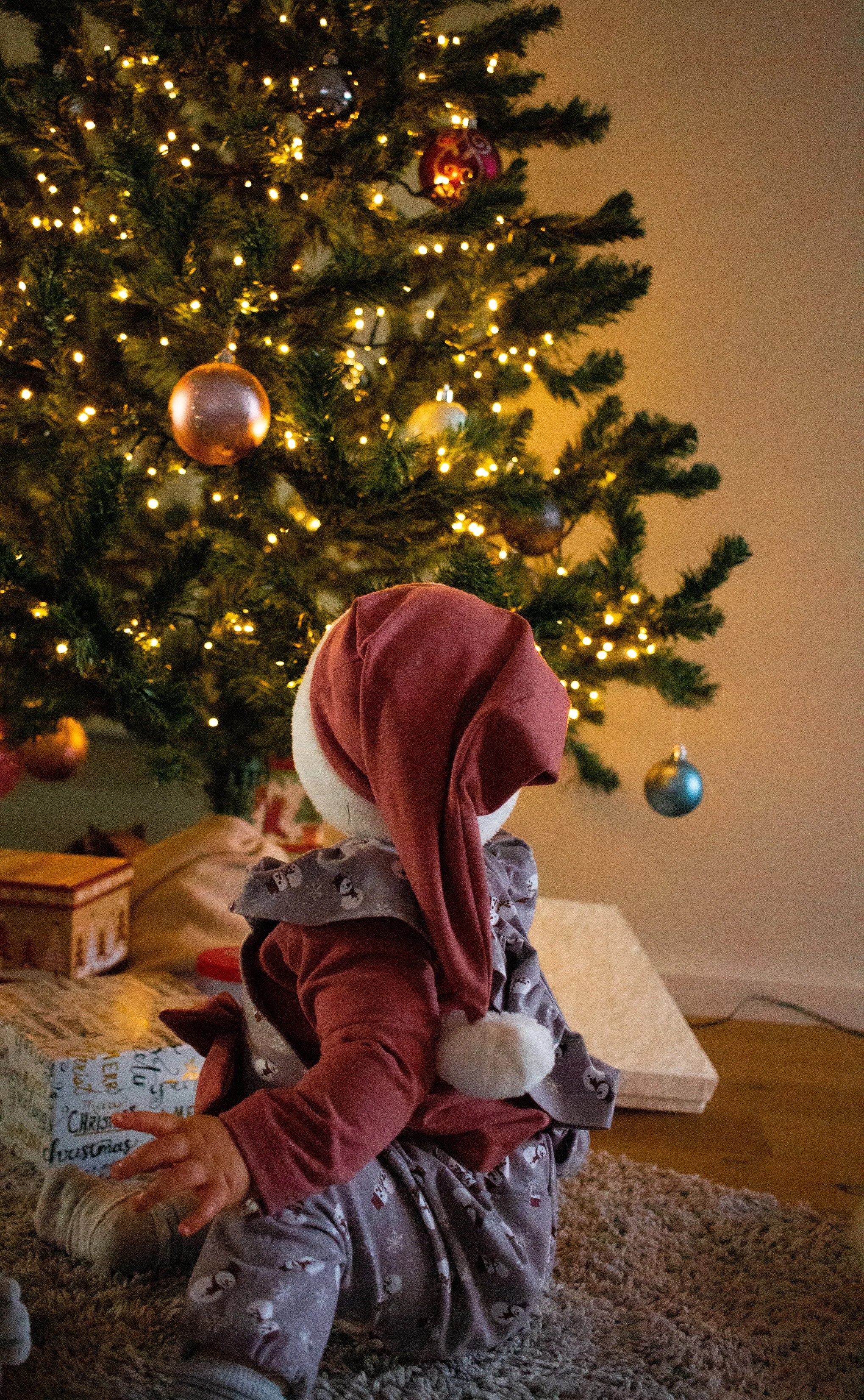 Kleines Kind in Weihnachtsoutfit sitzt vor geschmücktem Weihnachtbaum.