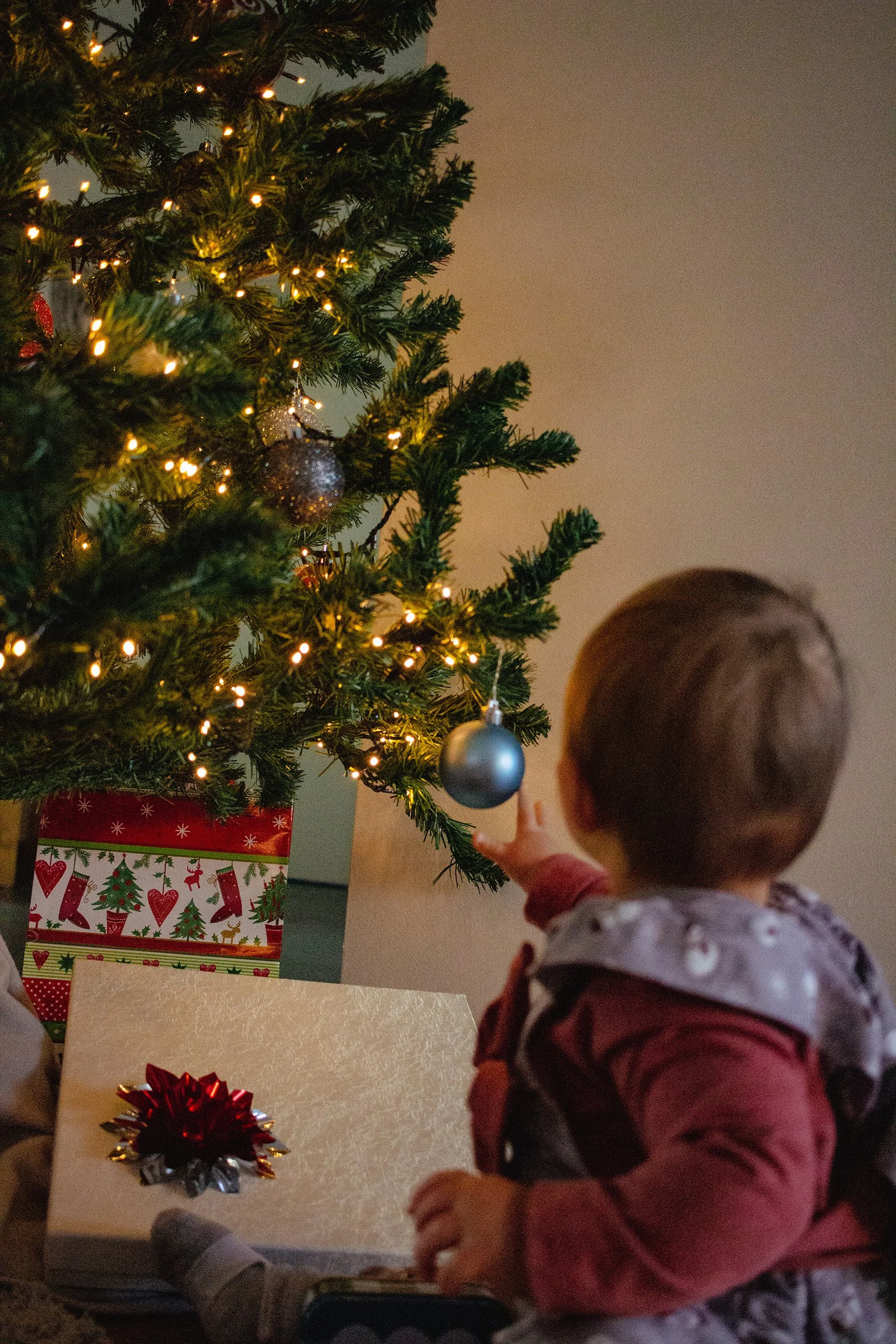 Ein kleines Kind schaut neugierig auf einen geschmückten Weihnachtsbaum mit Lichtern und Kugeln, im Hintergrund ein weihnachtlich verpacktes Geschenk.